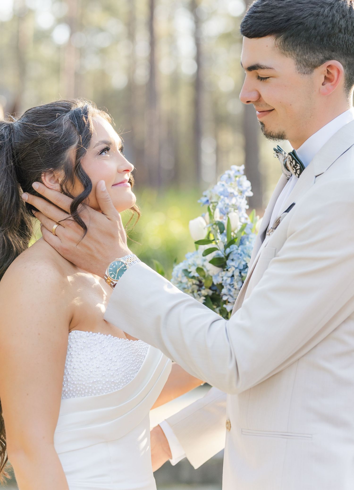 Groom in ivory suit tenderly cups bride's face during romantic outdoor first look wedding moment.