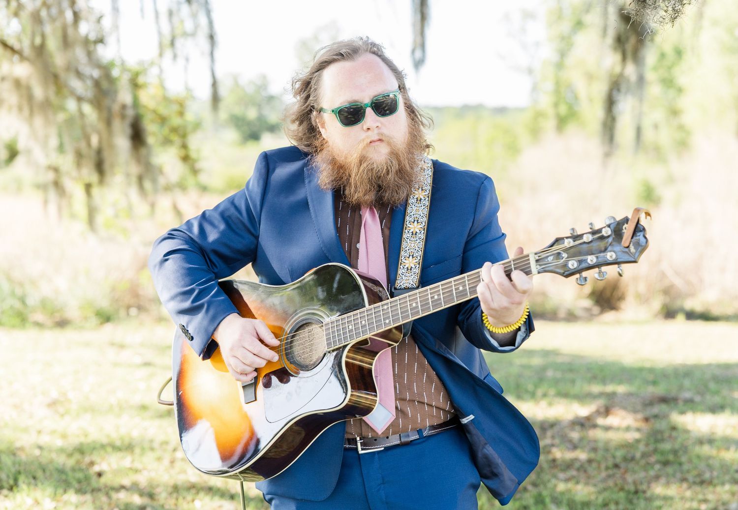 Bearded musician in a blue suit and sunglasses playing acoustic guitar outdoors at a wedding.