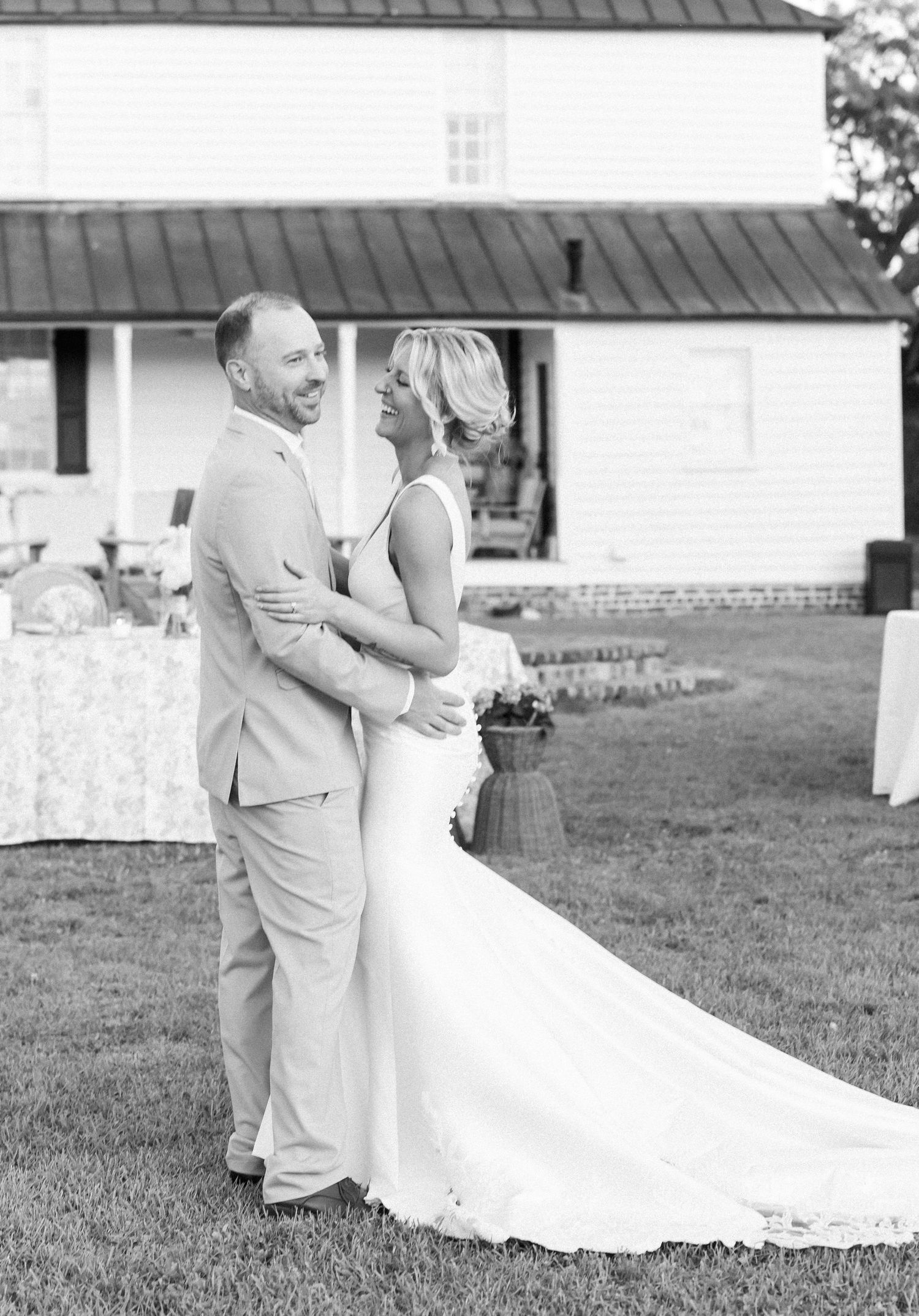 Black and white photo of smiling bride and groom embracing outdoors near farmhouse reception venue.