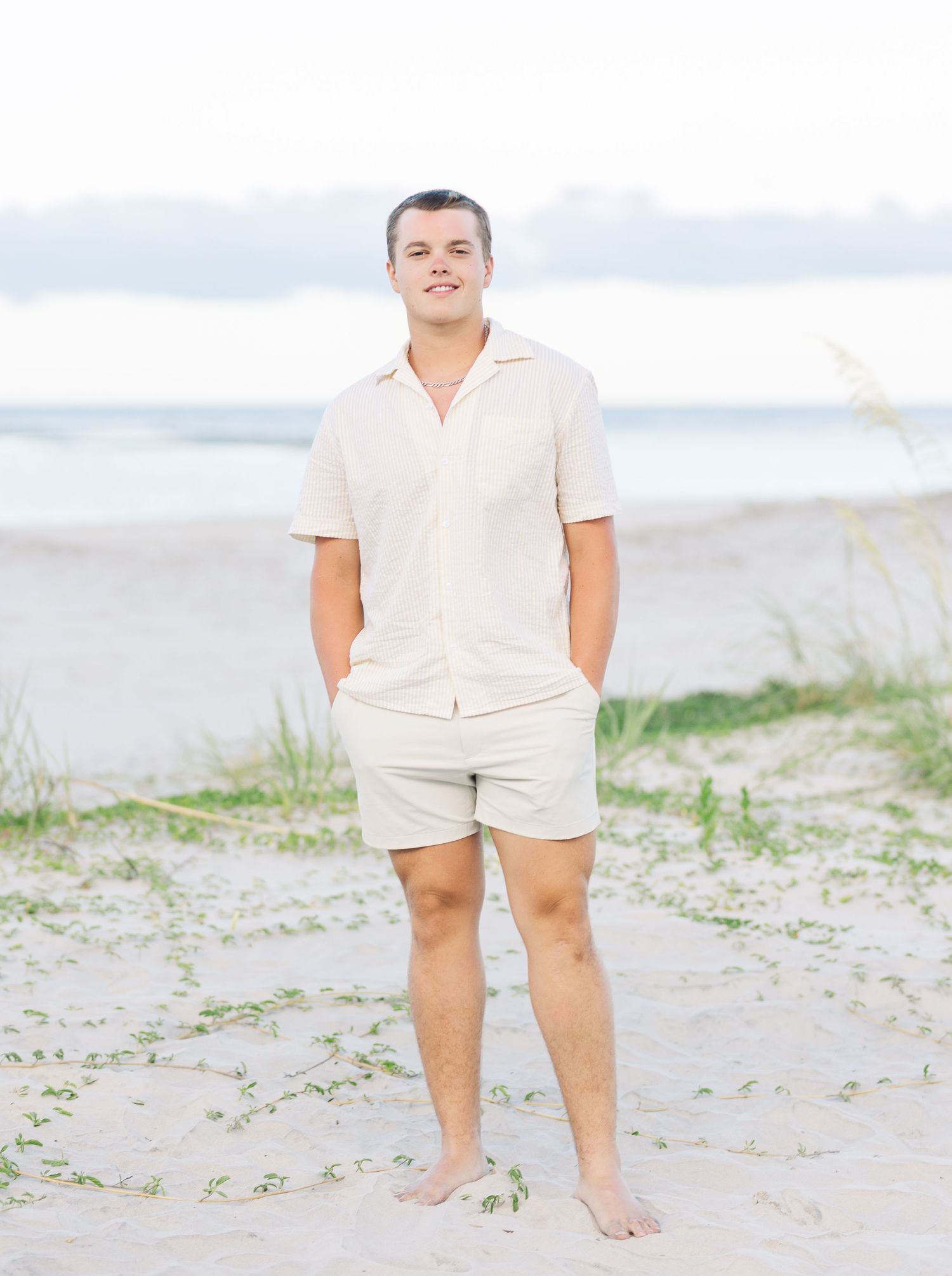 A person in a beige polo and shorts stands barefoot on a beach.