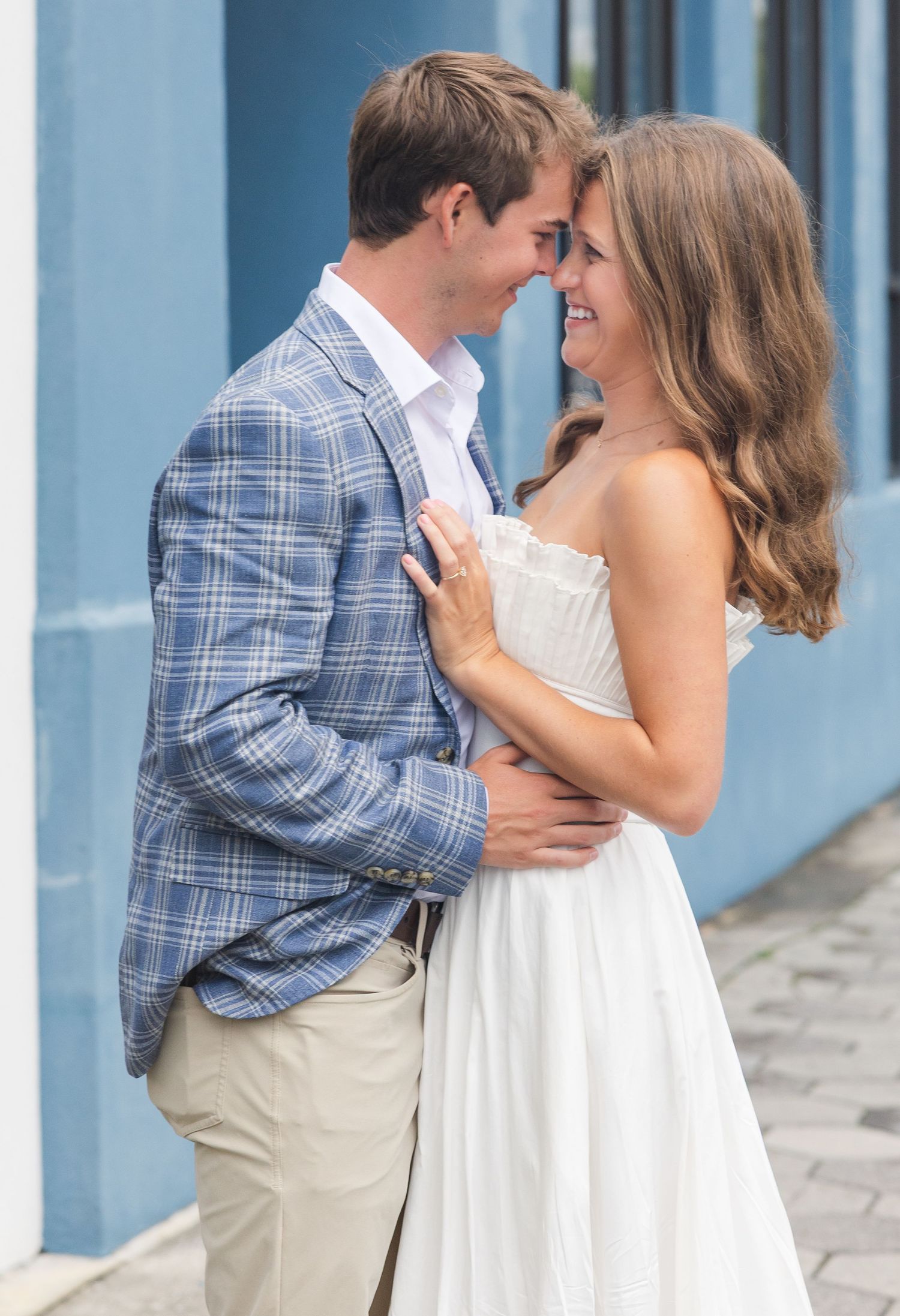Couple in romantic embrace, man in plaid blazer and woman in white dress, smiling at each other outdoors.