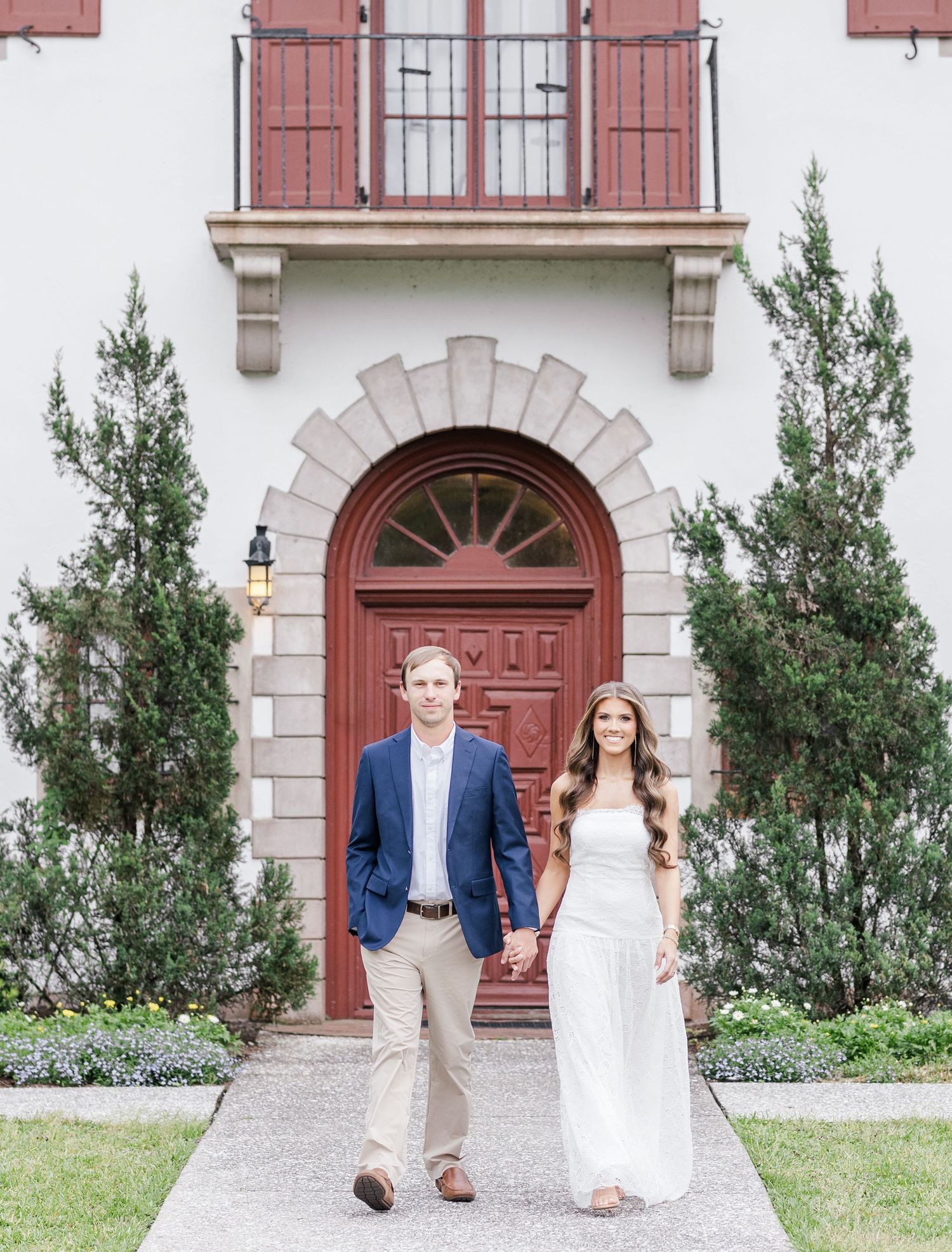 Couple in wedding attire holding hands in front of elegant building with red arched door and cypress trees.