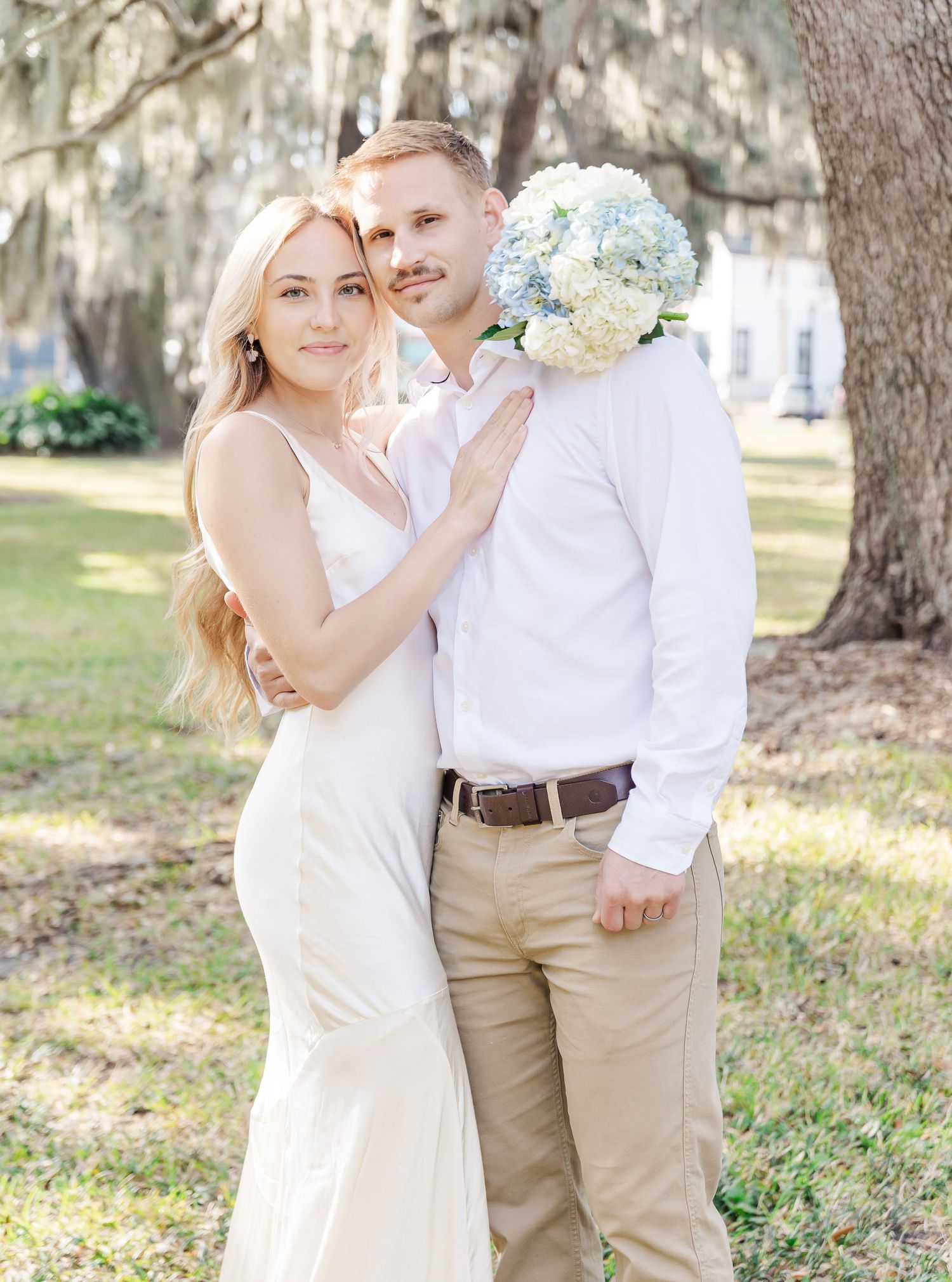 An elegant couple poses with light blue hydrangea bouquet under moss-draped oak trees.