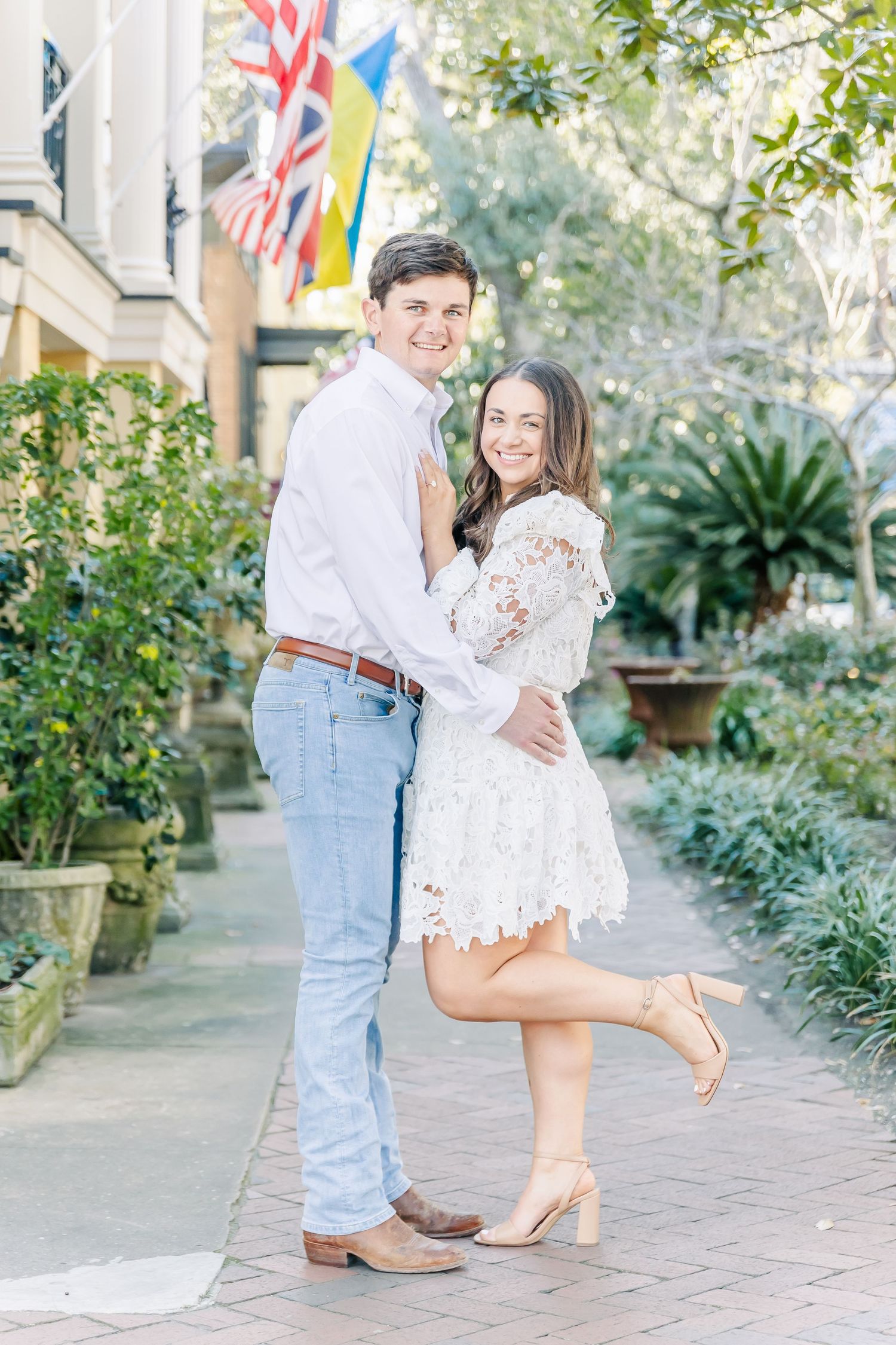 Couple in coordinating white outfits embracing and smiling on a brick pathway with flags overhead.