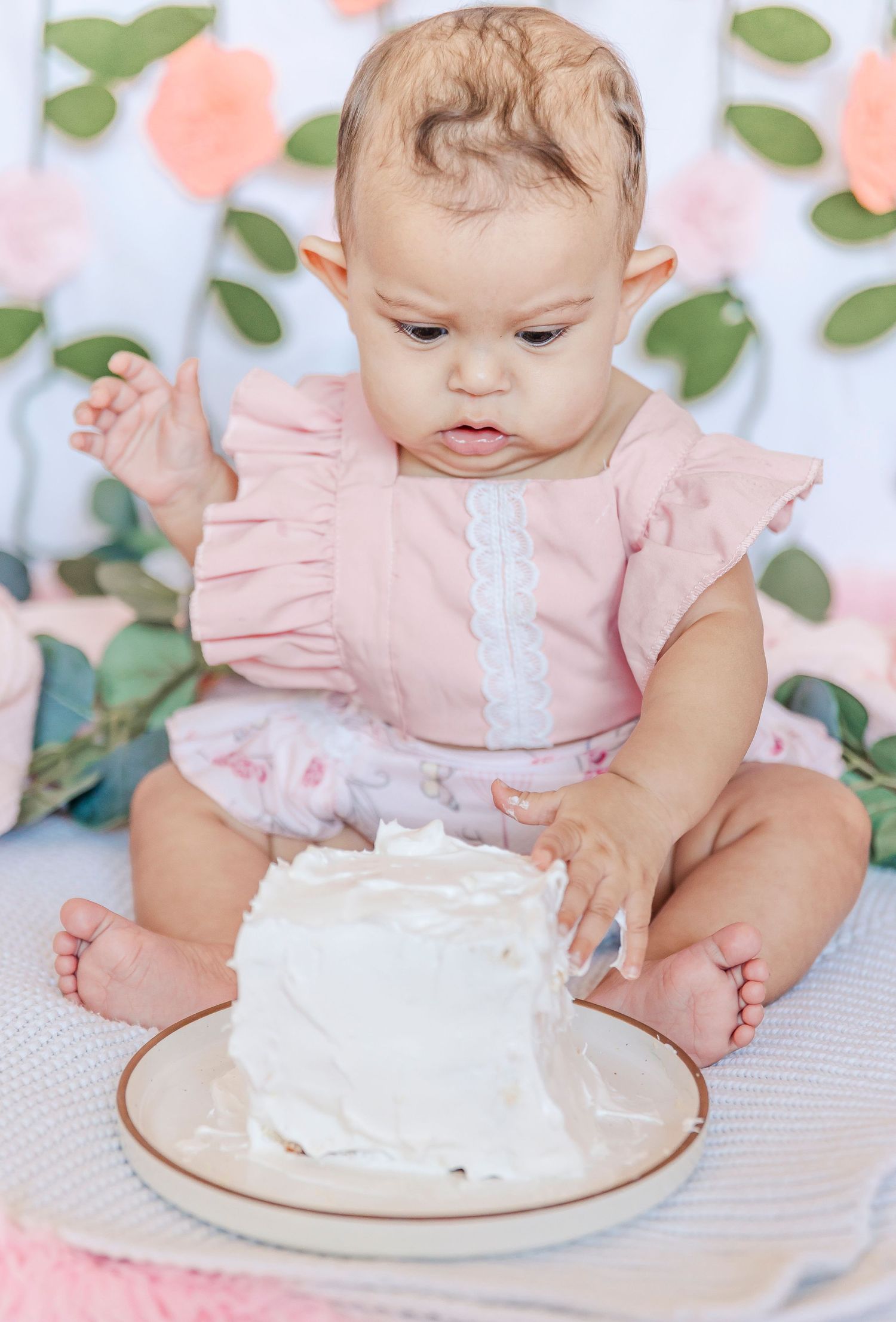 Baby in pink outfit exploring small white cake during photo session.