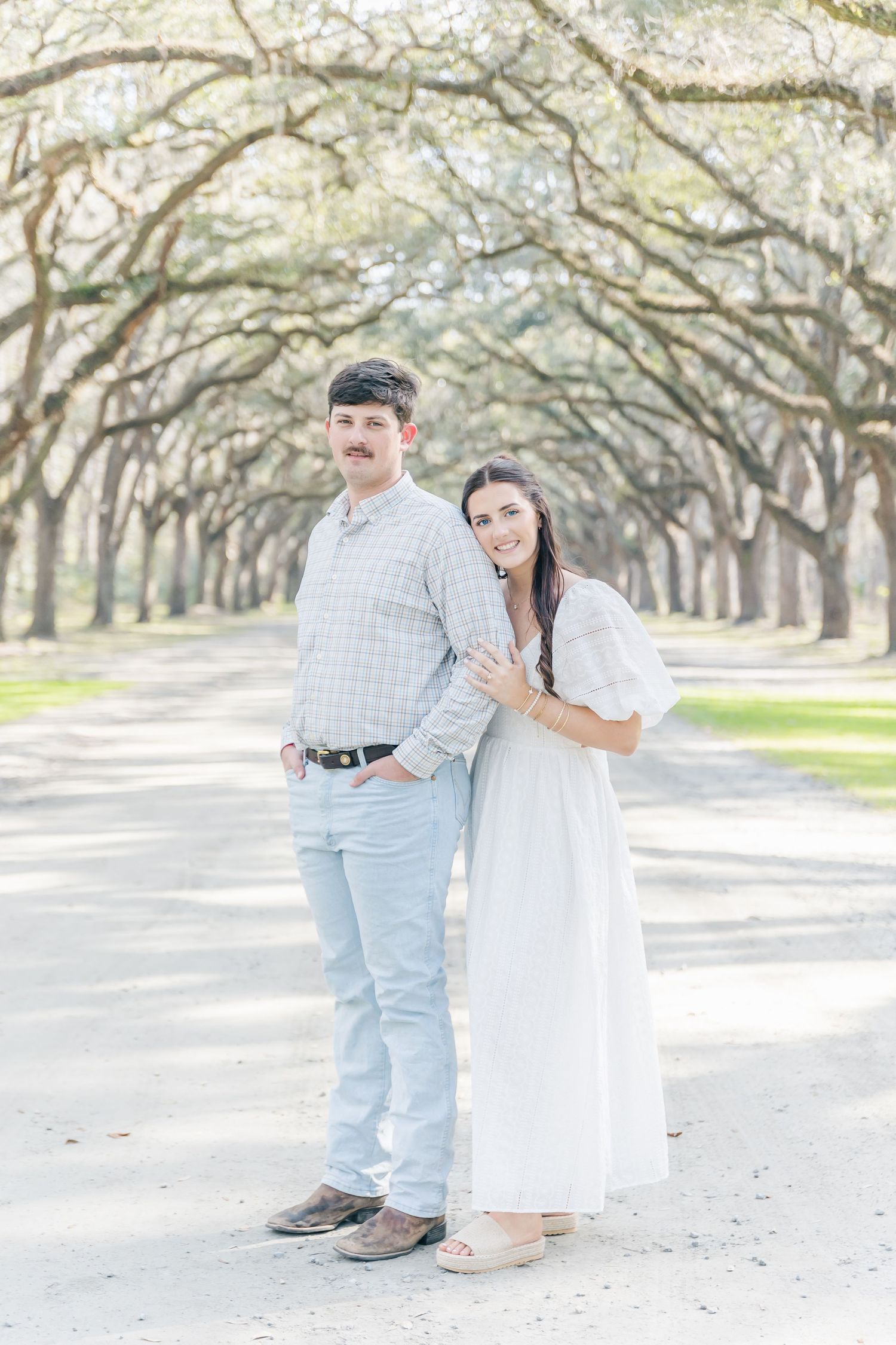 A couple poses on a tree-lined path wearing light colored outfits in a romantic outdoor setting.