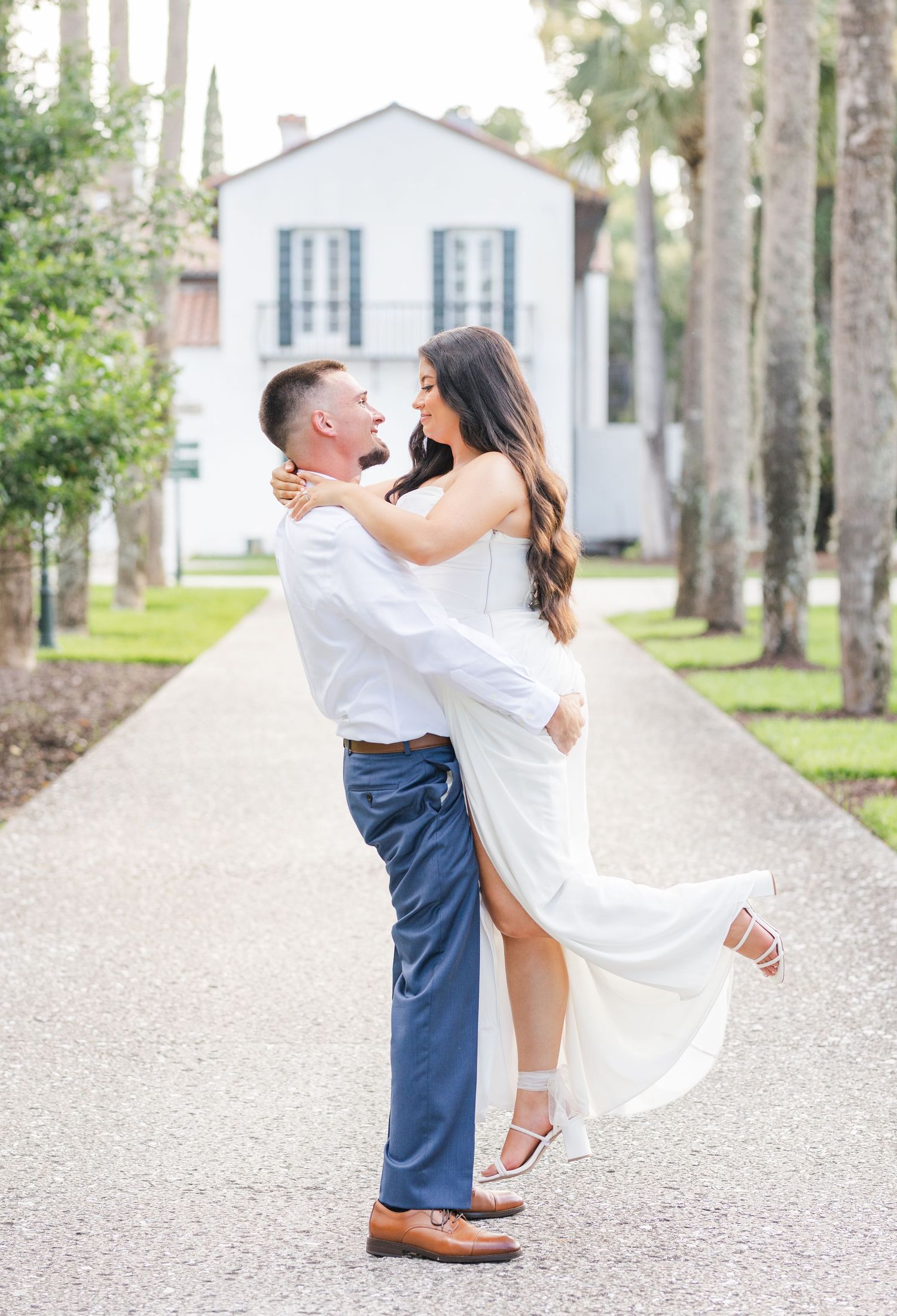 A groom lifts his bride in a white dress on a tree-lined path, with a white estate building in the background.