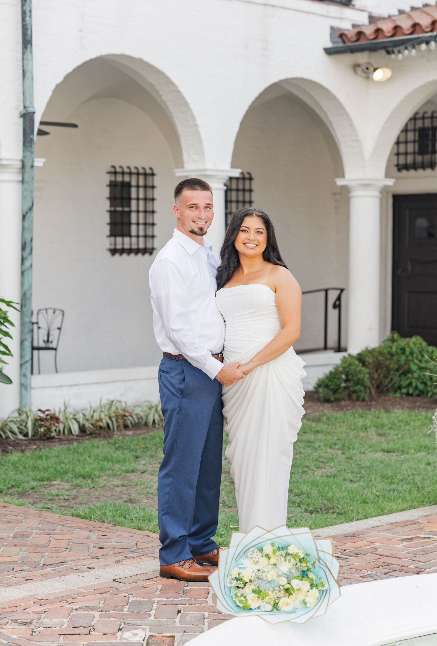 Couple posing in front of white arched building, woman in white dress holding bouquet, man in blue pants.