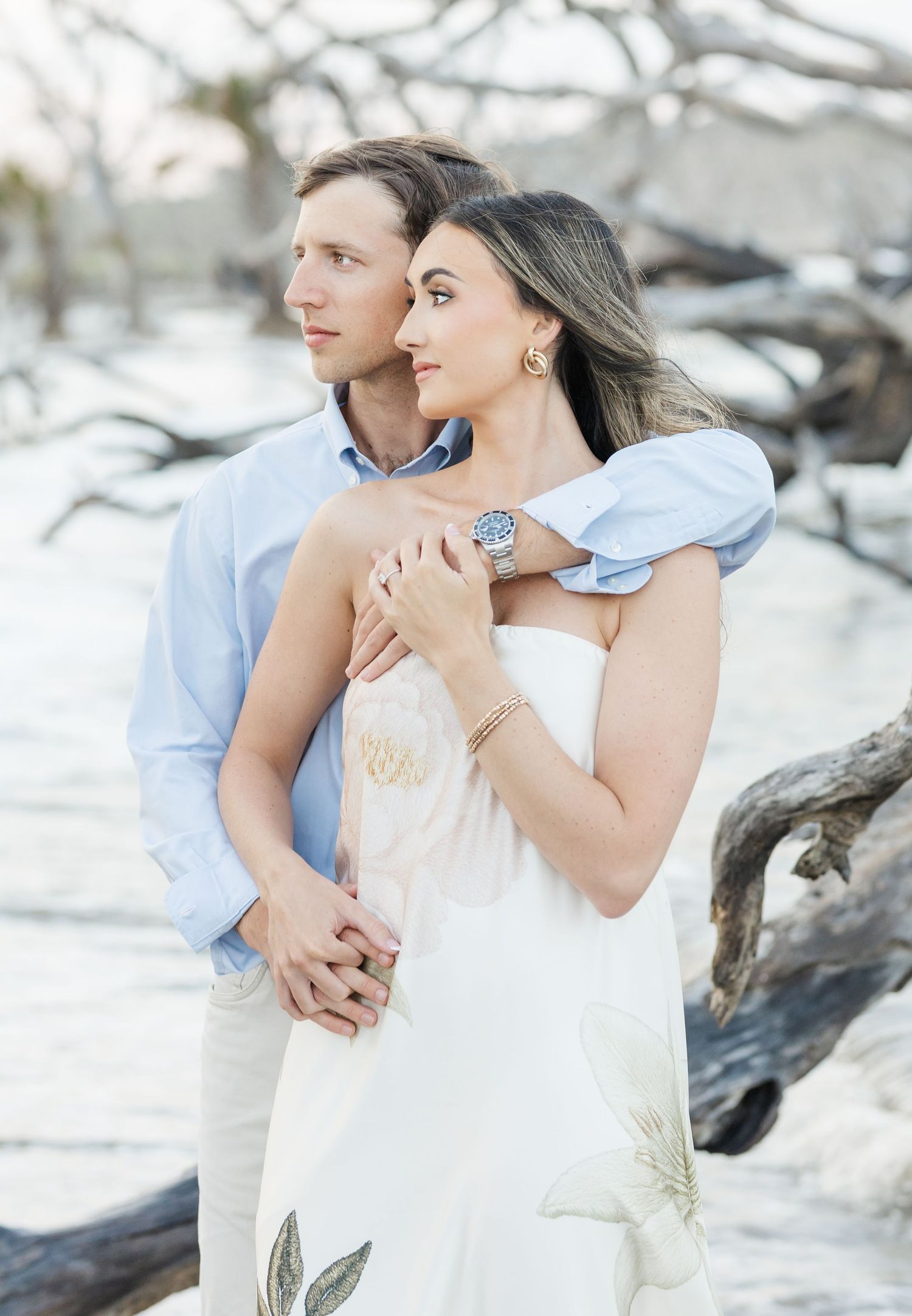 Elegant couple embracing near driftwood on beach, woman wearing floral white dress and Rolex watch.