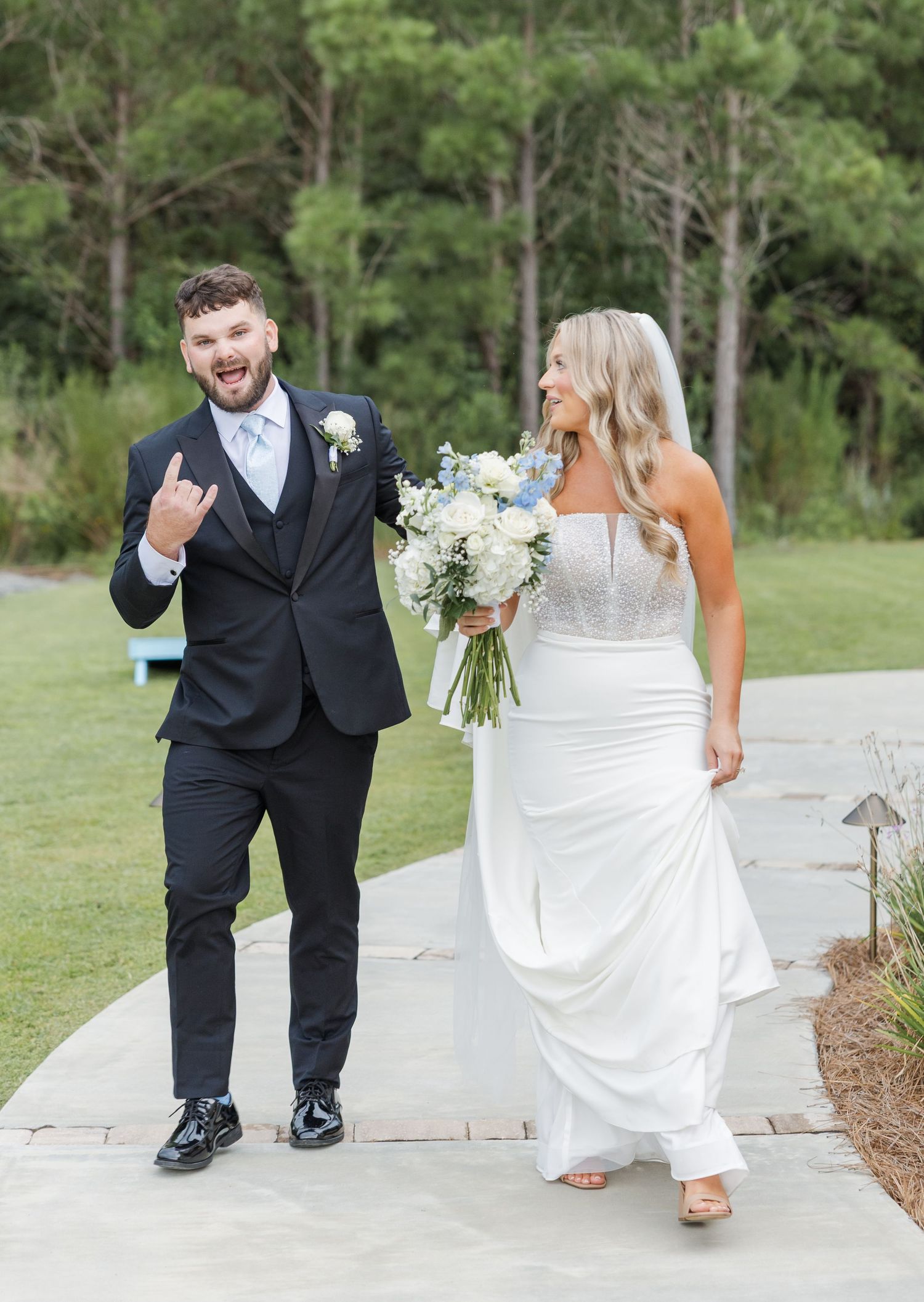 Joyful newlyweds walking together after wedding ceremony, groom in black suit making rock sign, bride holding bouquet.