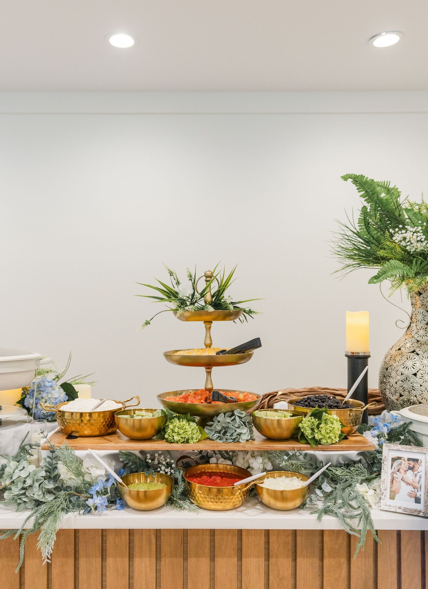 Gold serving bowls and tiered stand on a wooden buffet table decorated with greenery and blue hydrangeas.