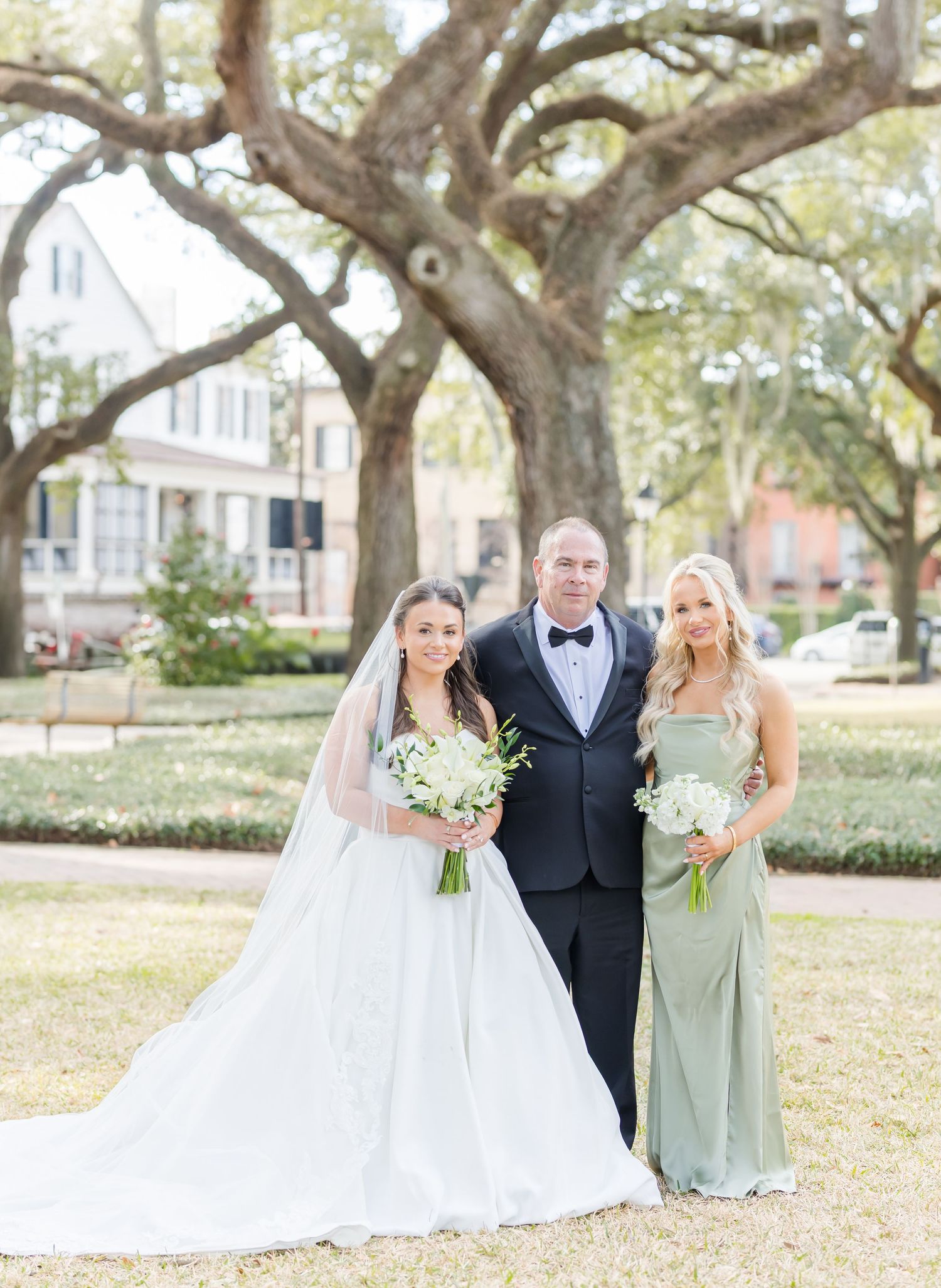 Bride in white ball gown with veil, groom in tuxedo, and bridesmaid in sage green dress posing outdoors.