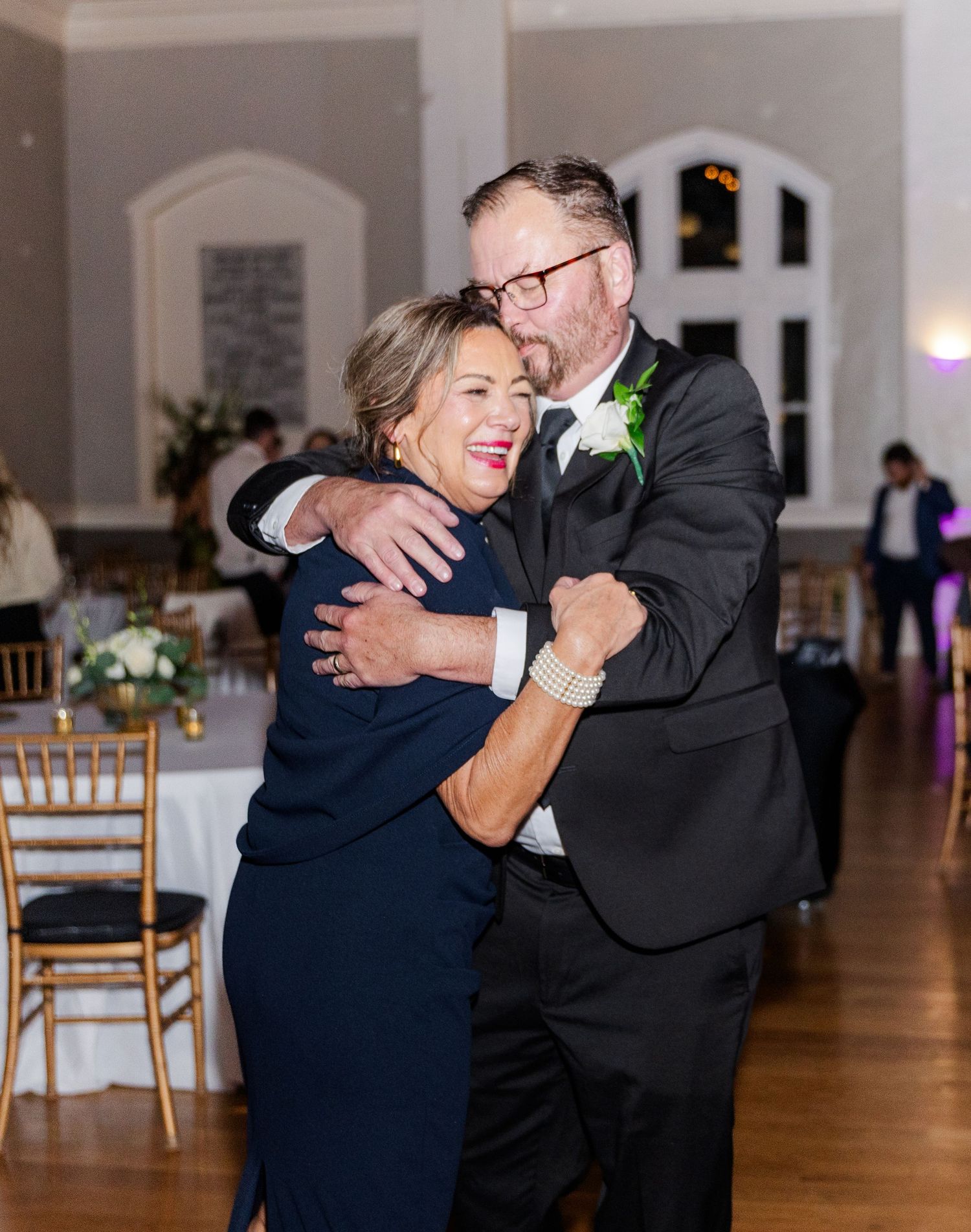 Groom in black tuxedo shares a warm embrace while dancing with woman in navy dress at reception.