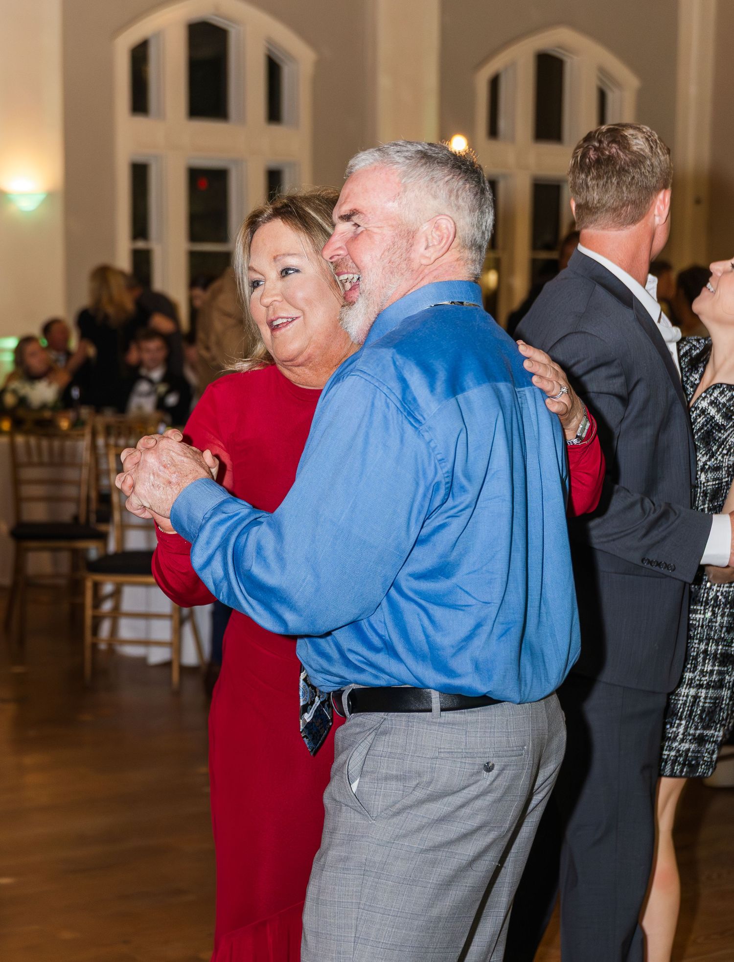 Couple dancing joyfully at wedding reception in ballroom, woman in red dress and man in blue shirt.