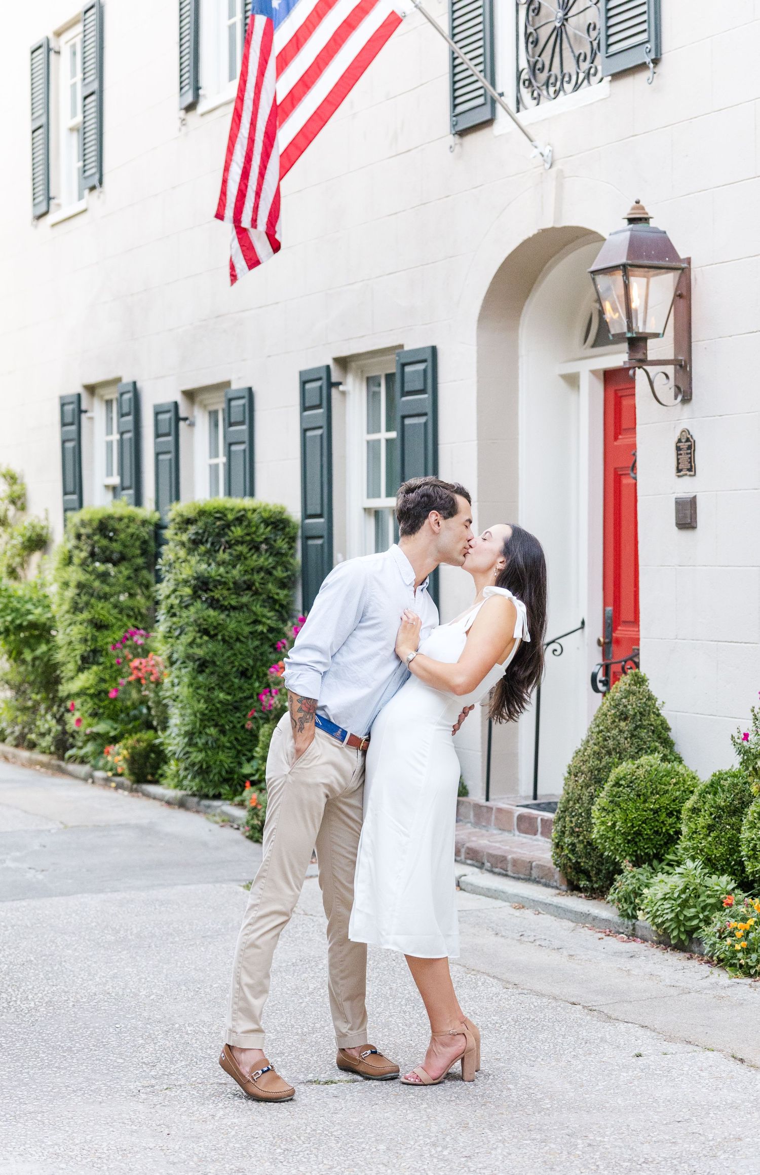 Couple kissing in front of a charming white building adorned with an American flag and green shutters.