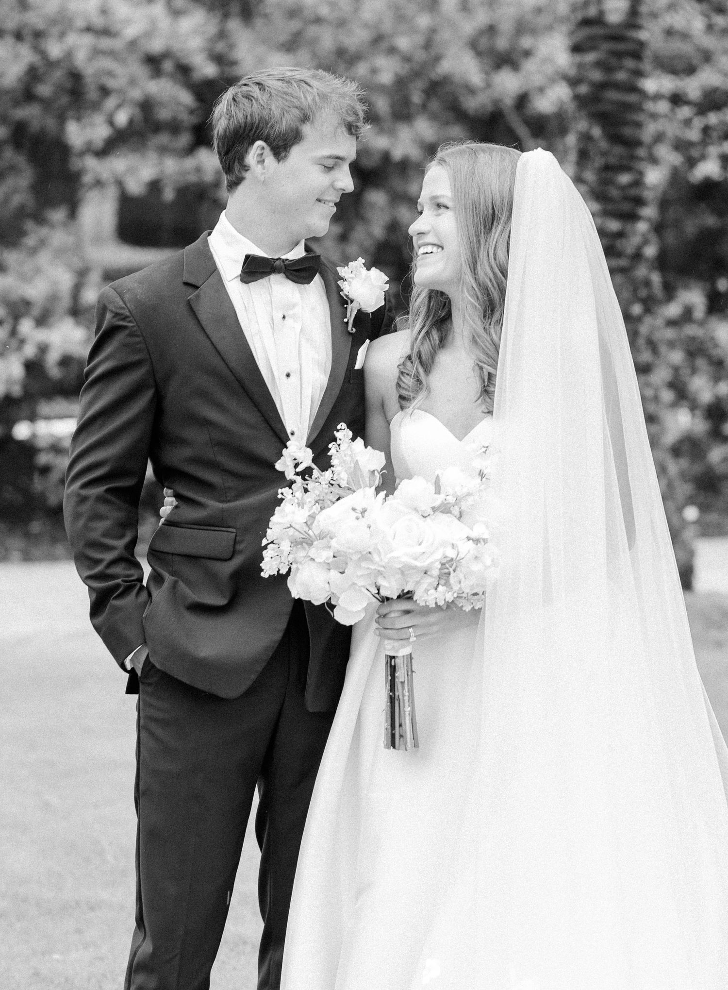 Black and white wedding portrait of smiling bride in veil holding bouquet and groom in tuxedo gazing at each other outdoors.