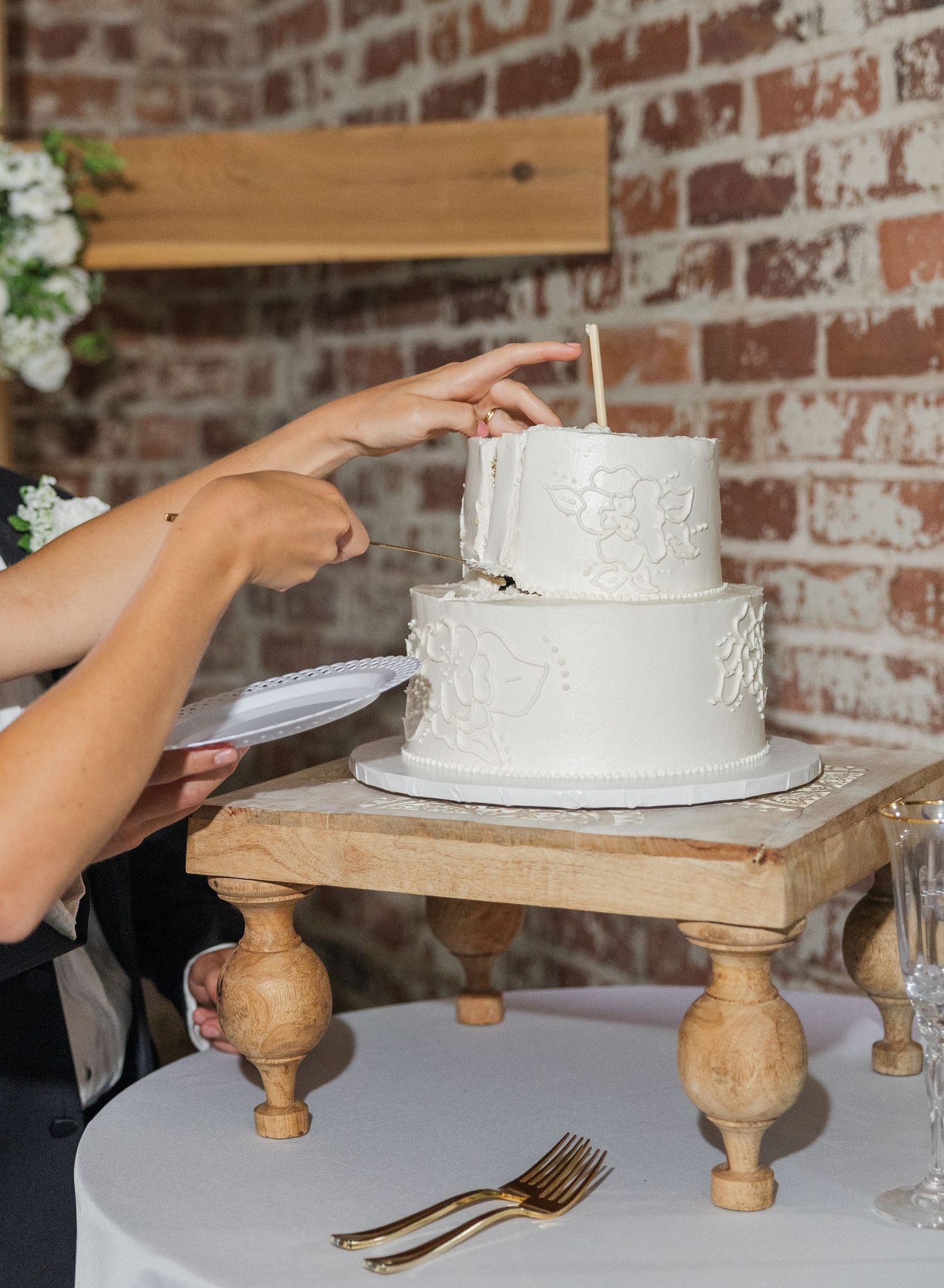 Bride and groom cut elegant two-tier white wedding cake with floral details on rustic wooden cake stand.