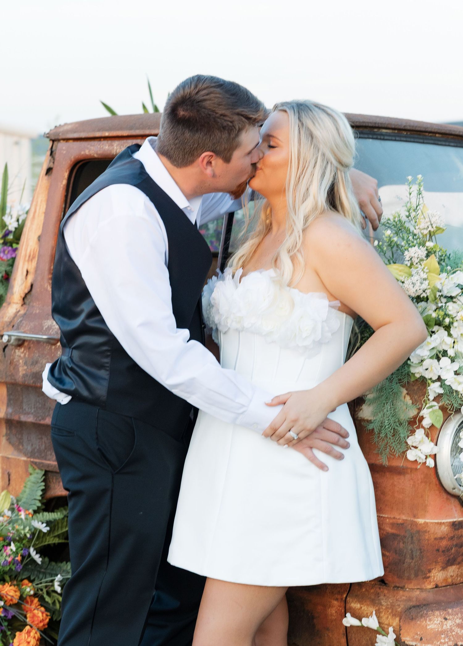 Bride and groom kissing in front of a rustic vintage truck decorated with colorful flowers.