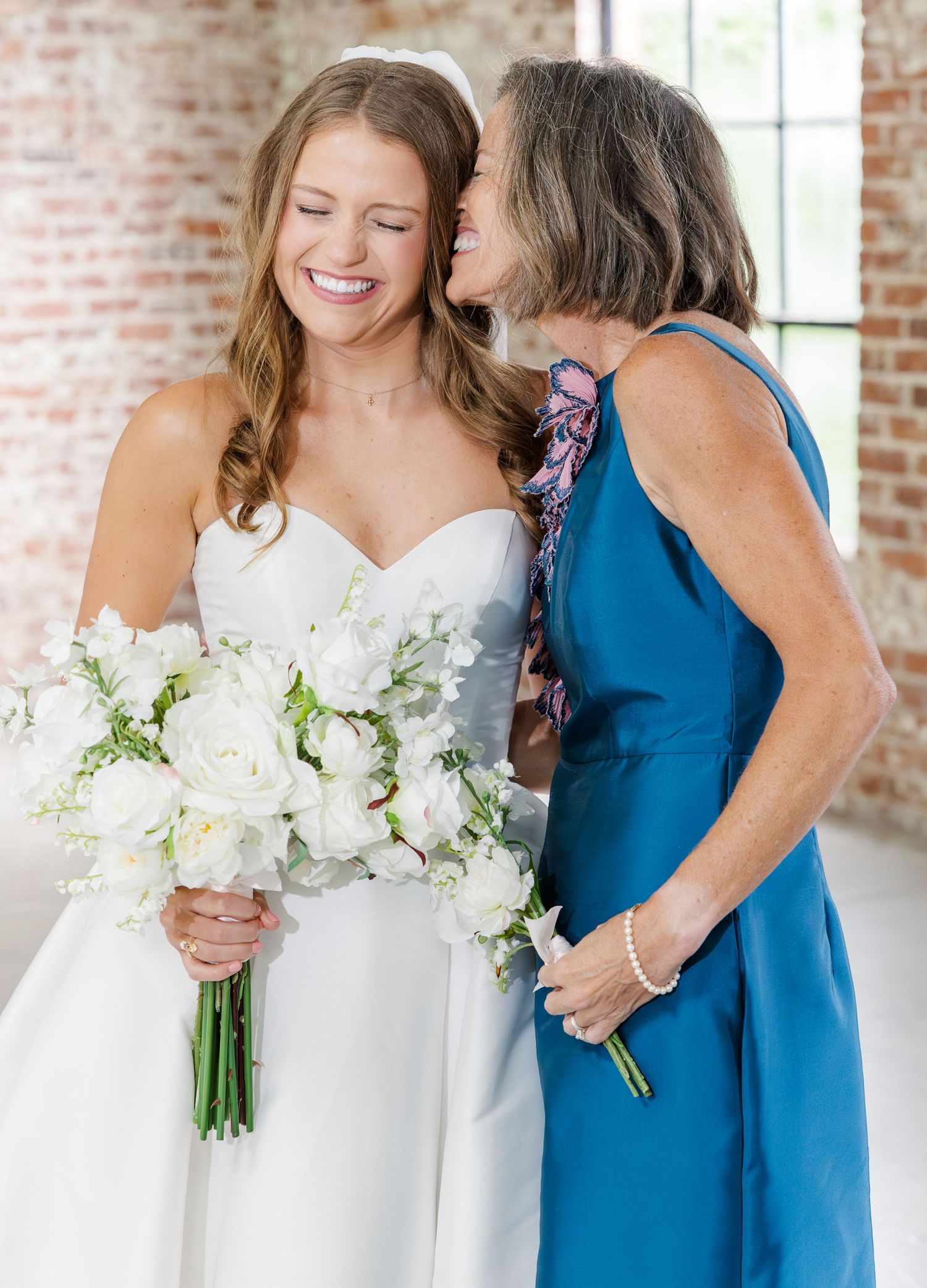Mother whispers to laughing bride in white sweetheart gown holding white floral bouquet indoors.