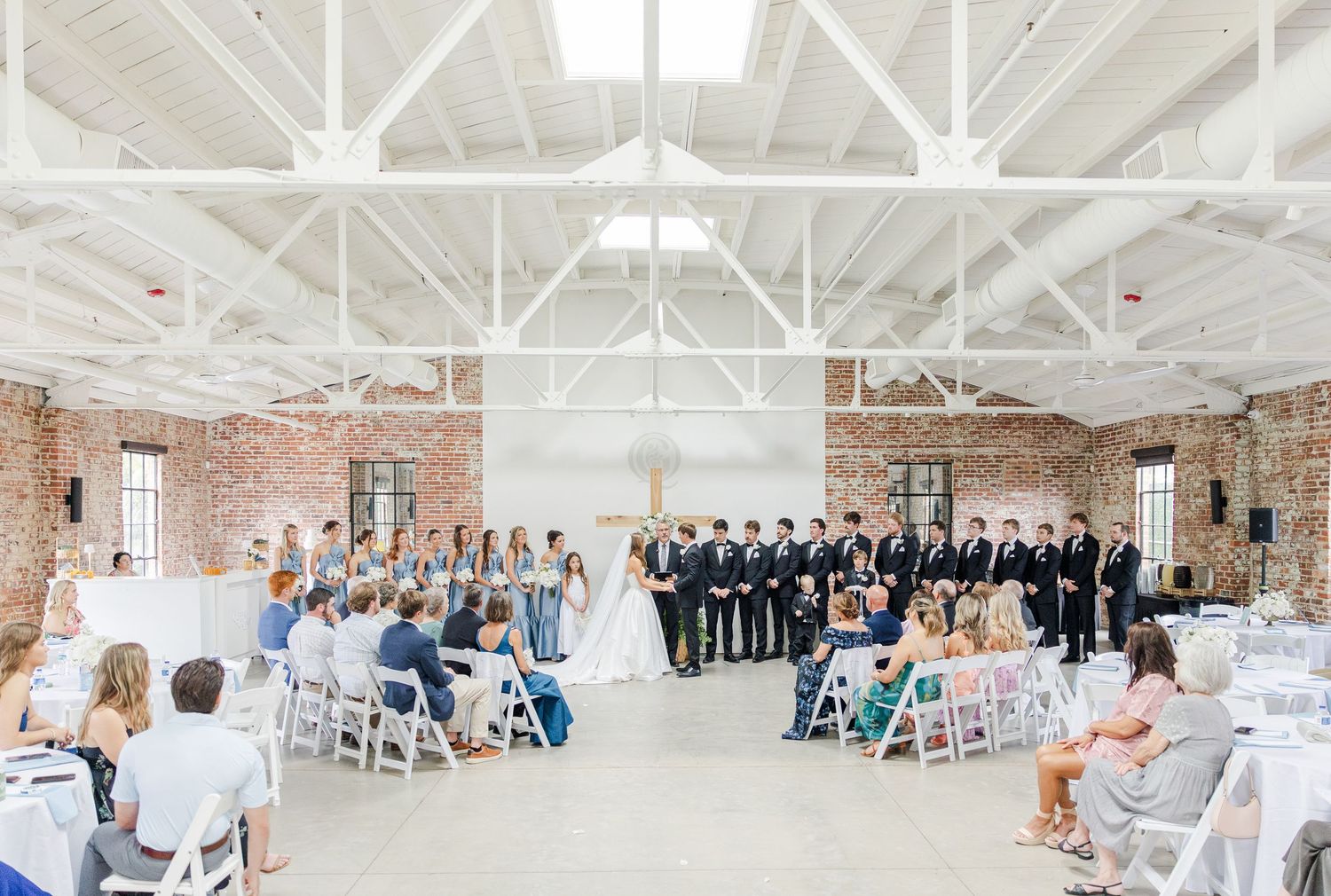 Wide angle view of elegant wedding ceremony in white industrial venue with brick walls and seated guests.