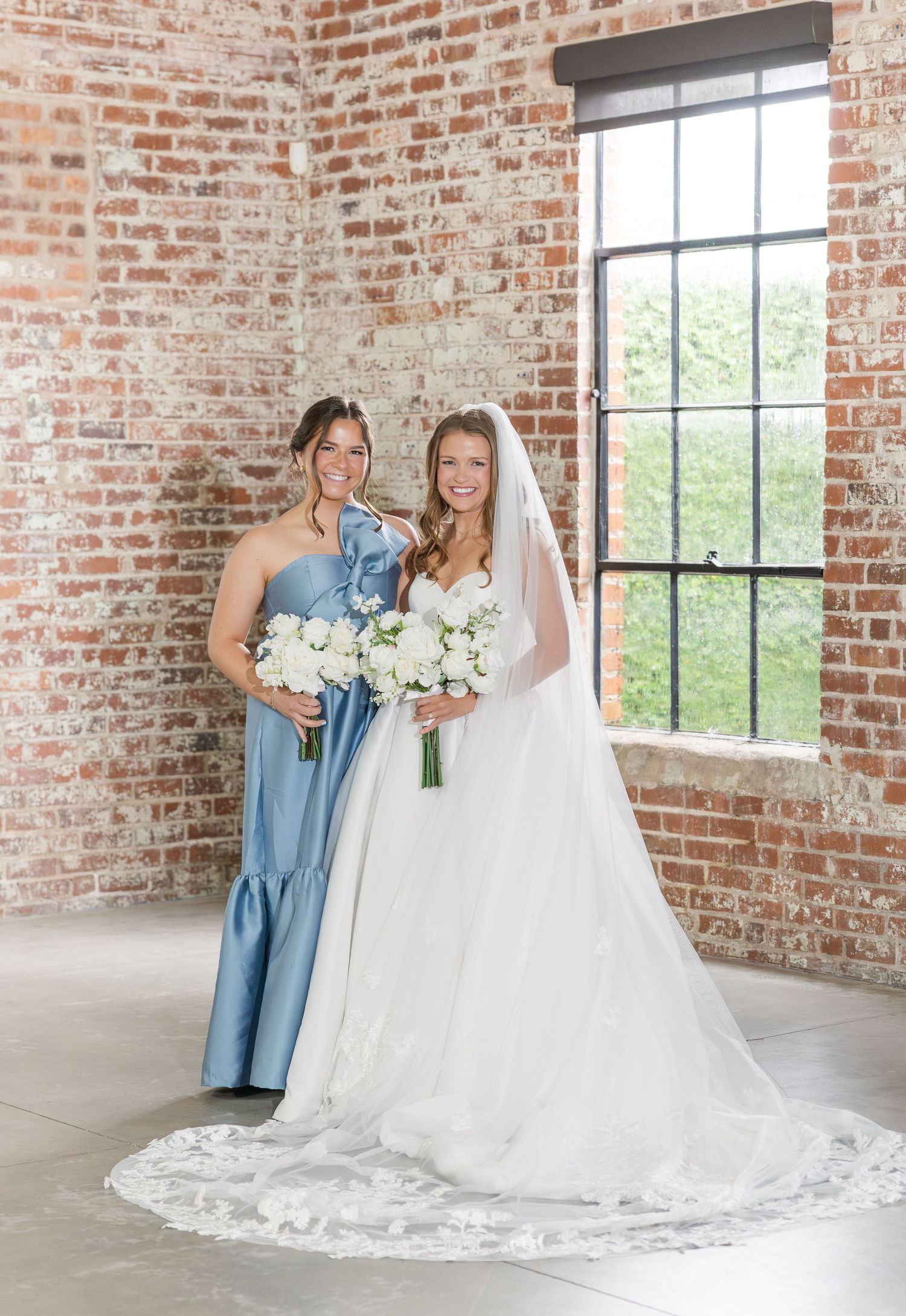 Bride in white gown with veil and bridesmaid in blue dress posing near brick wall and window.