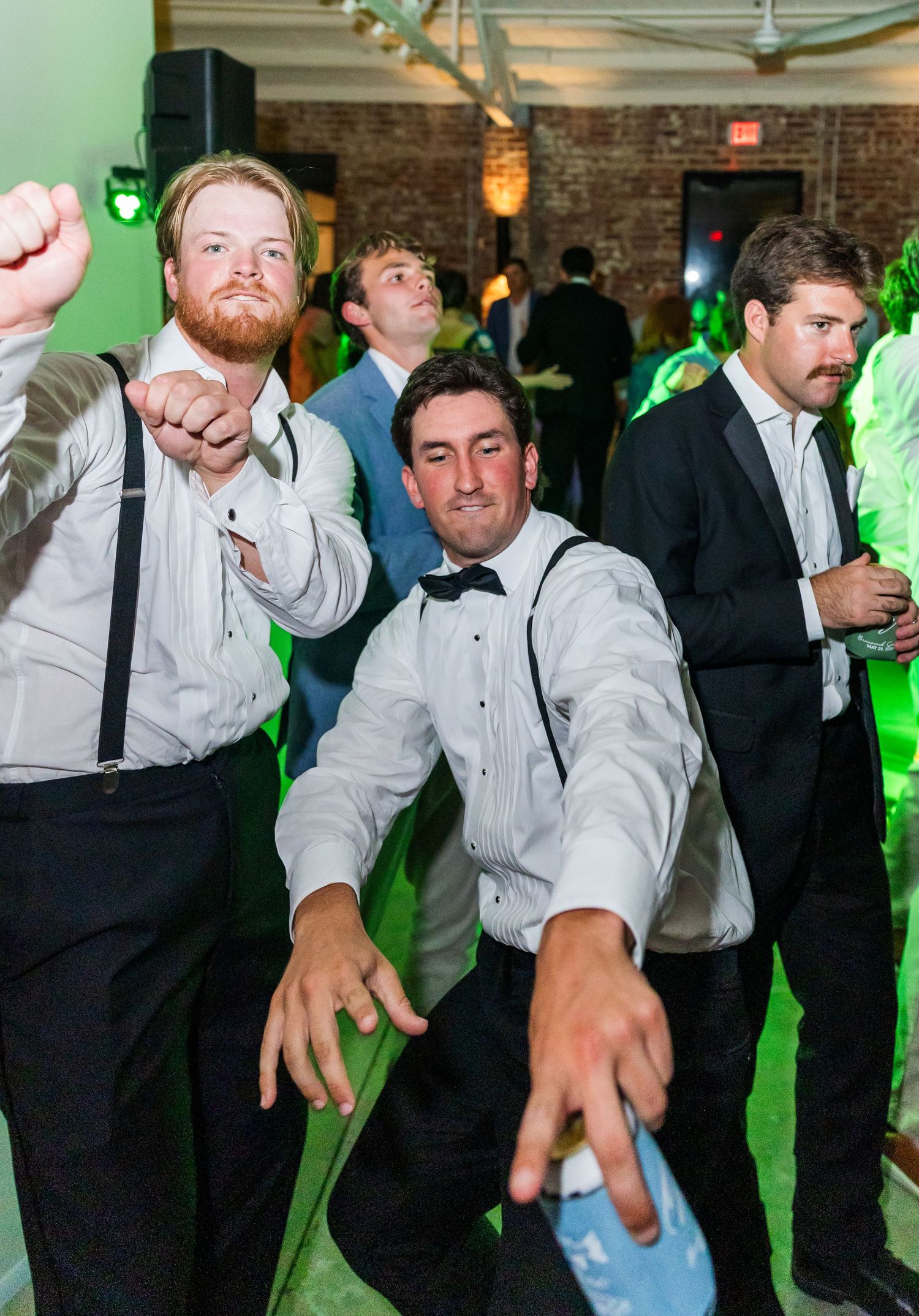 Groomsmen in white shirts and suspenders strike fun poses on green-lit wedding reception dance floor.