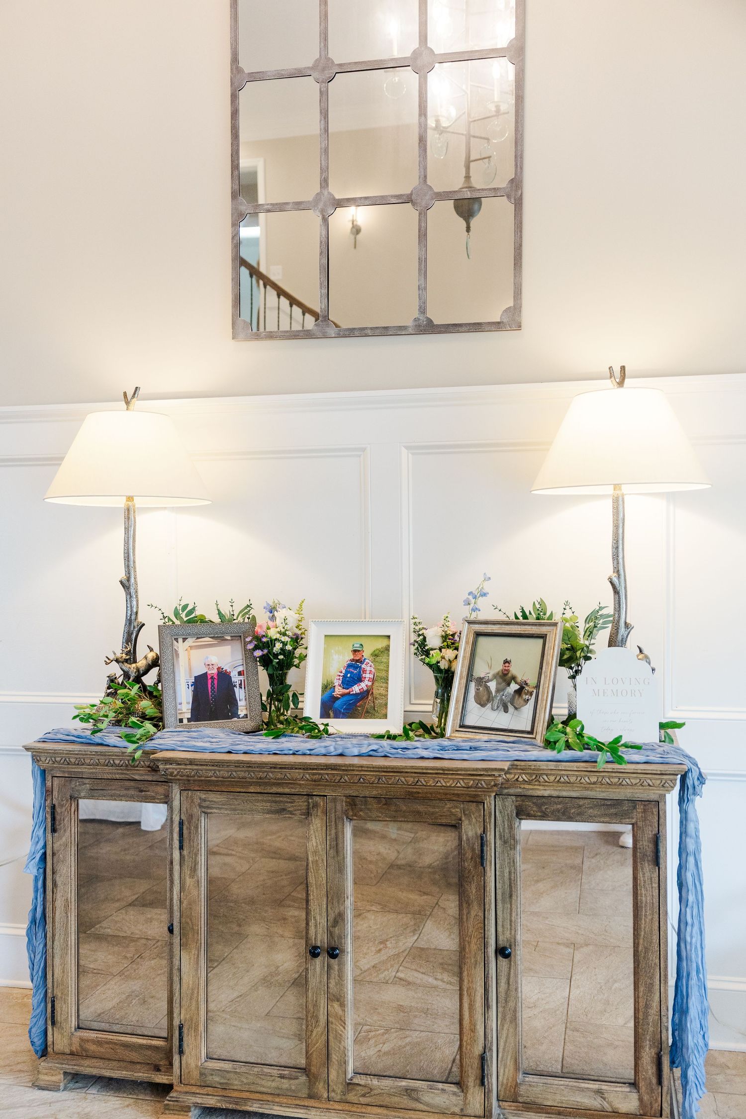 Rustic mirrored sideboard with framed photos, greenery, and white lamps beneath a large mirror.