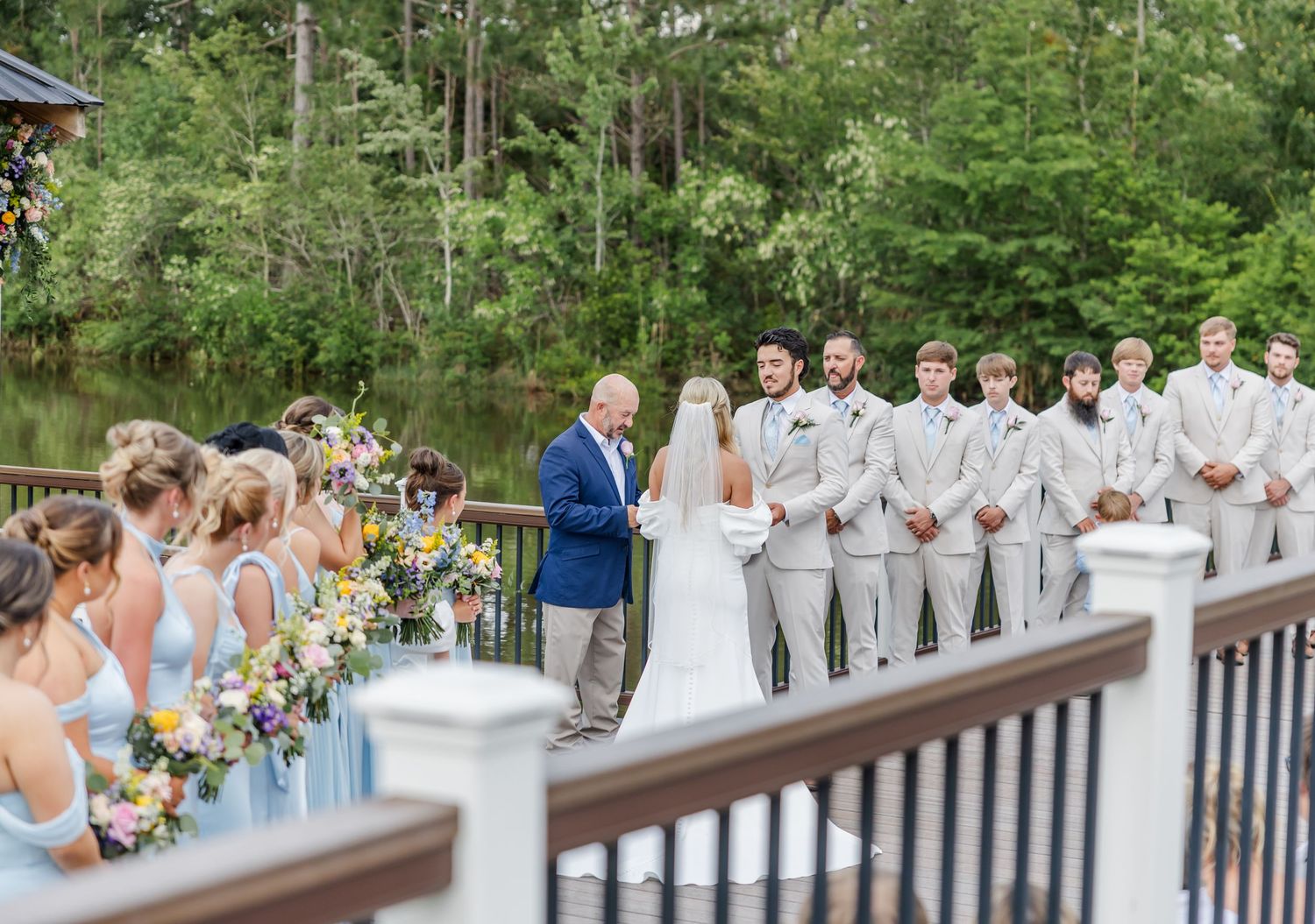 Wedding ceremony on waterfront deck with bridesmaids in blue dresses and groomsmen in gray suits.