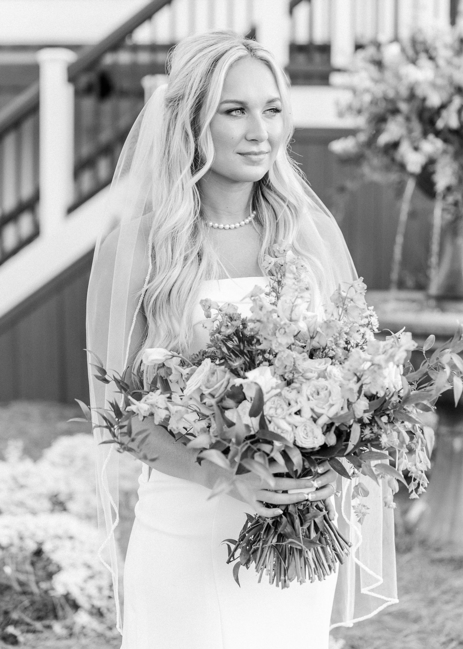 Black and white portrait of a bride in a white gown holding a floral bouquet with a veil and pearl necklace.