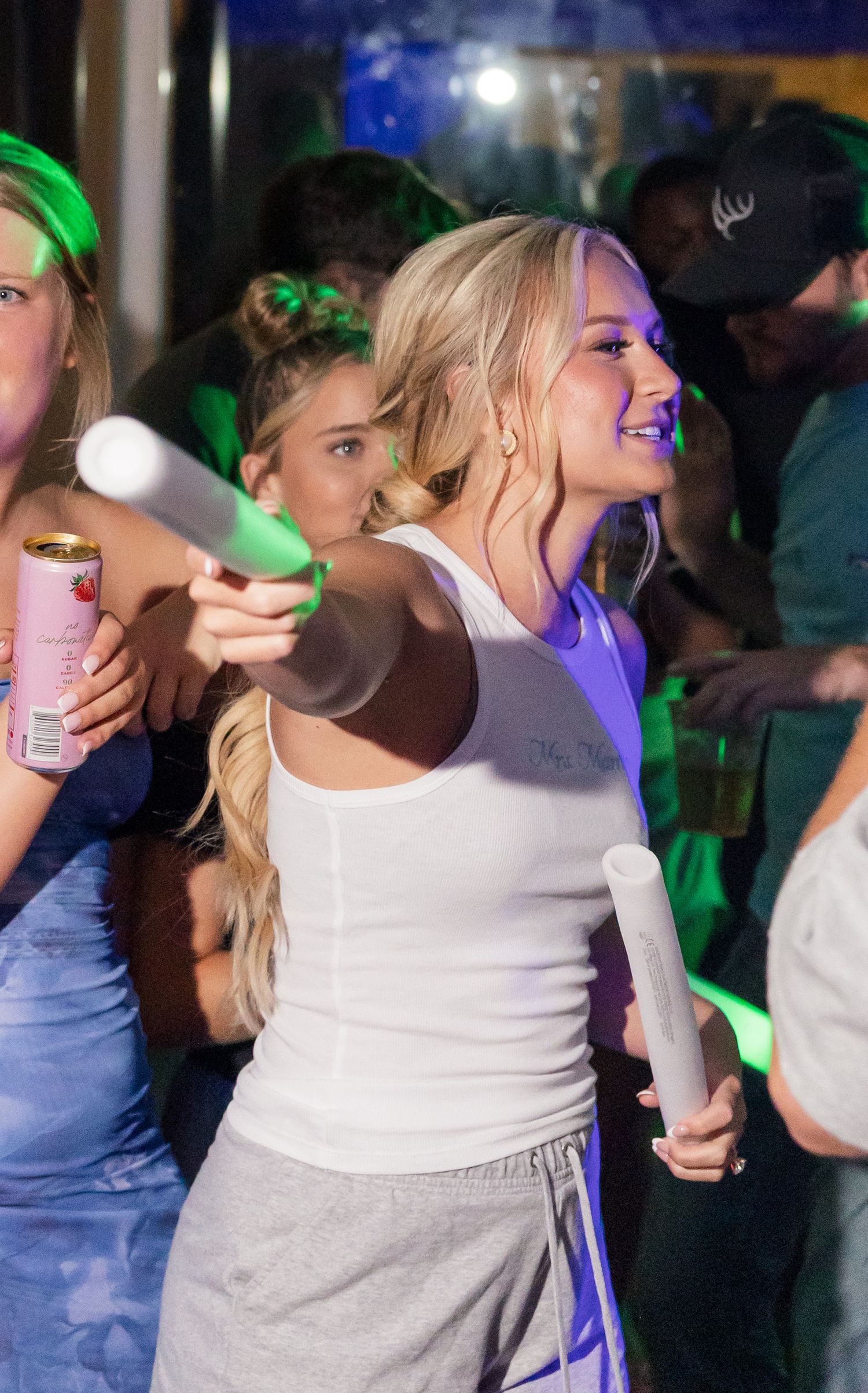 Blonde woman in white tank top celebrates at lively wedding reception with glow sticks.