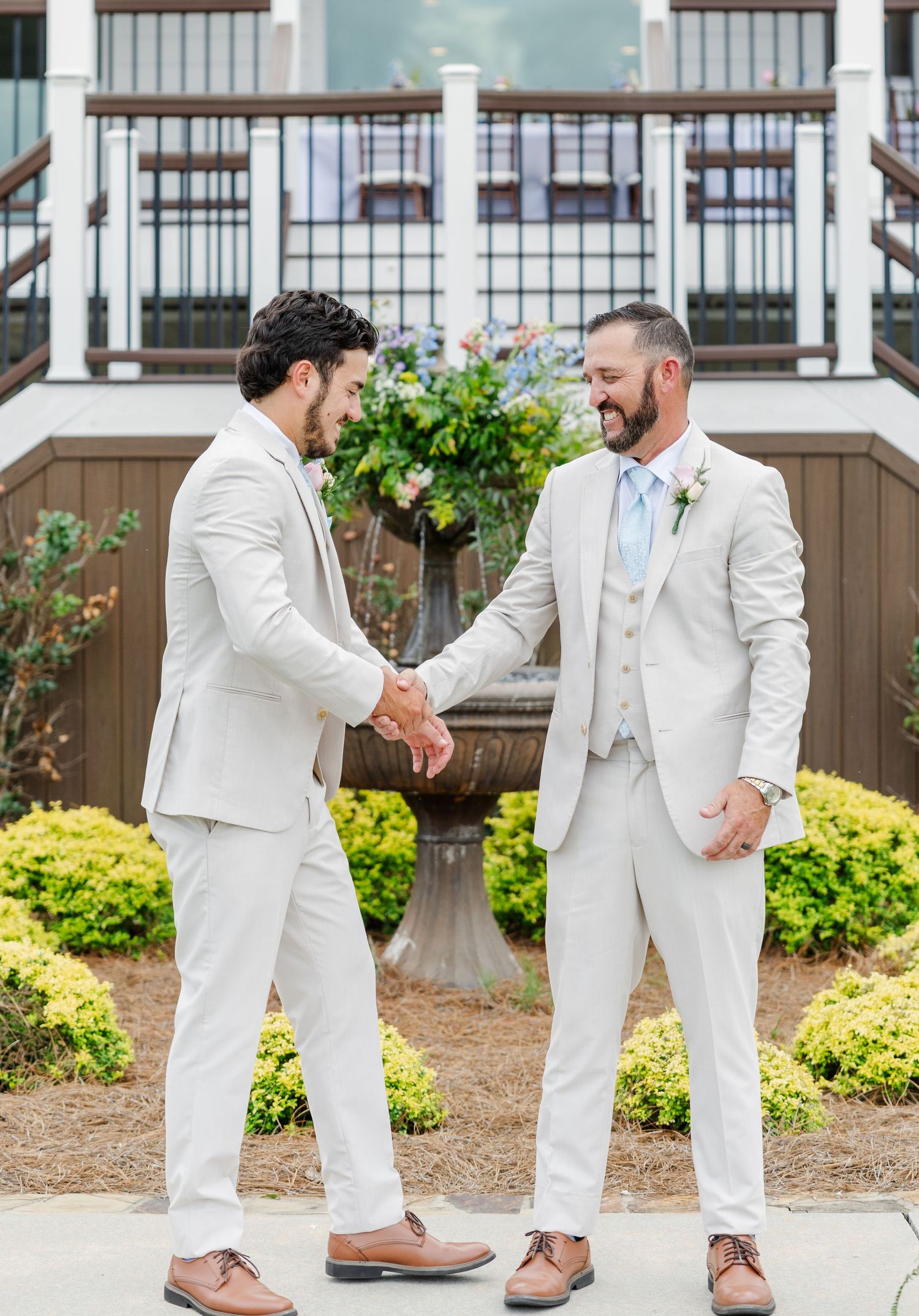 Two groomsmen in matching beige suits shake hands and smile outside a wedding venue with a fountain backdrop.