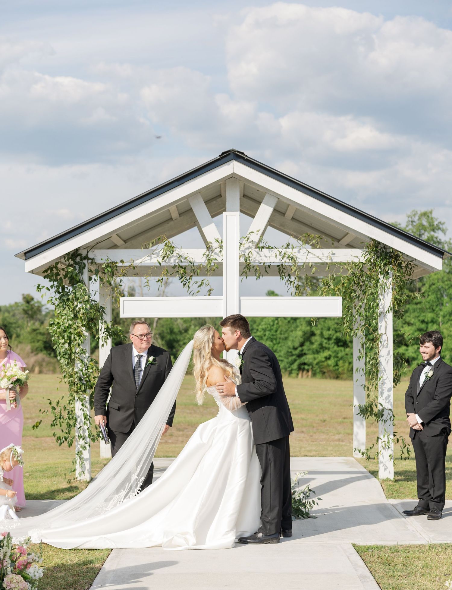 Bride and groom share first kiss under white pergola adorned with greenery at outdoor wedding ceremony.