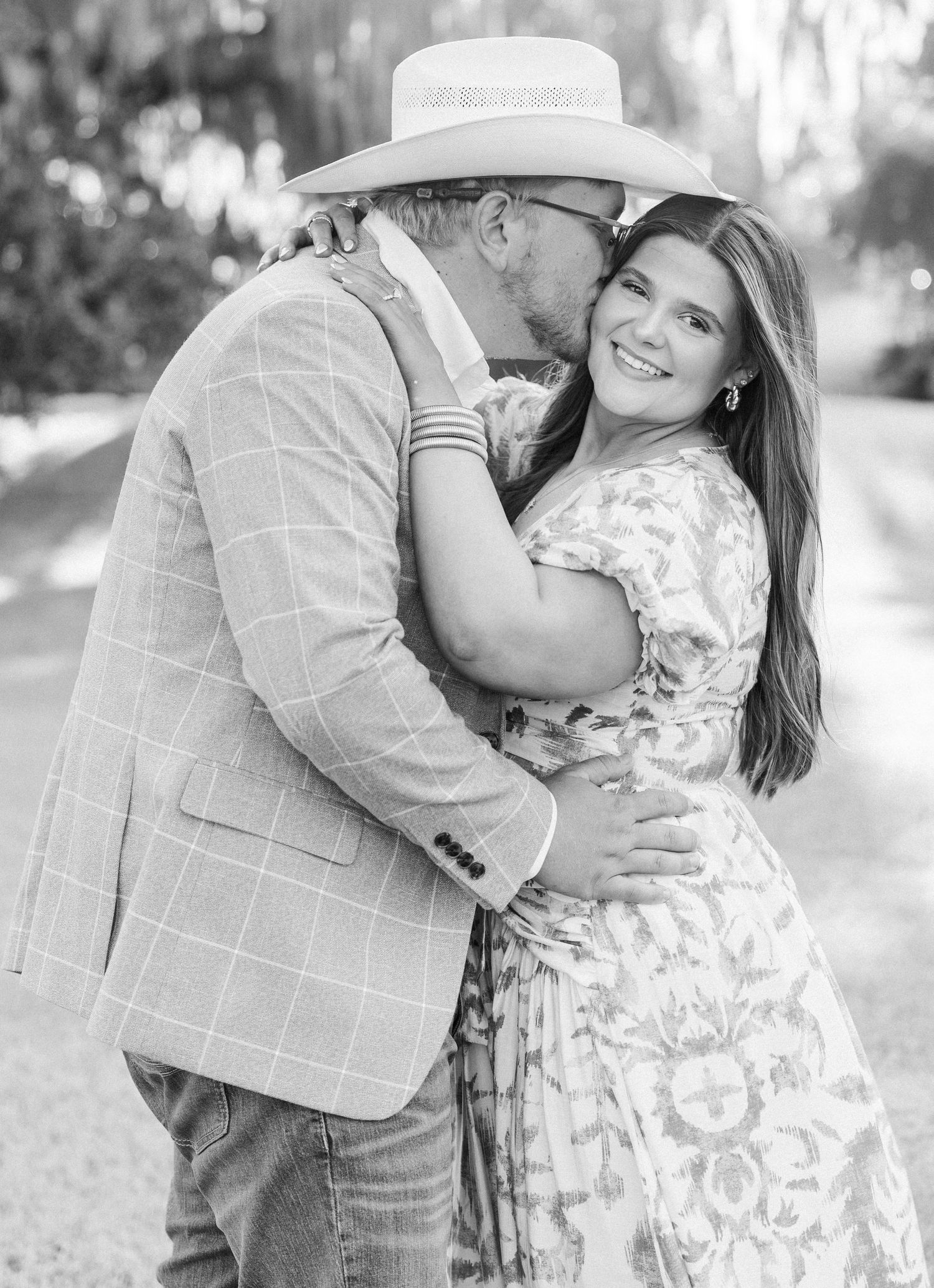 Black and white photo of a couple embracing outdoors, man in plaid blazer and cowboy hat kissing womans cheek.