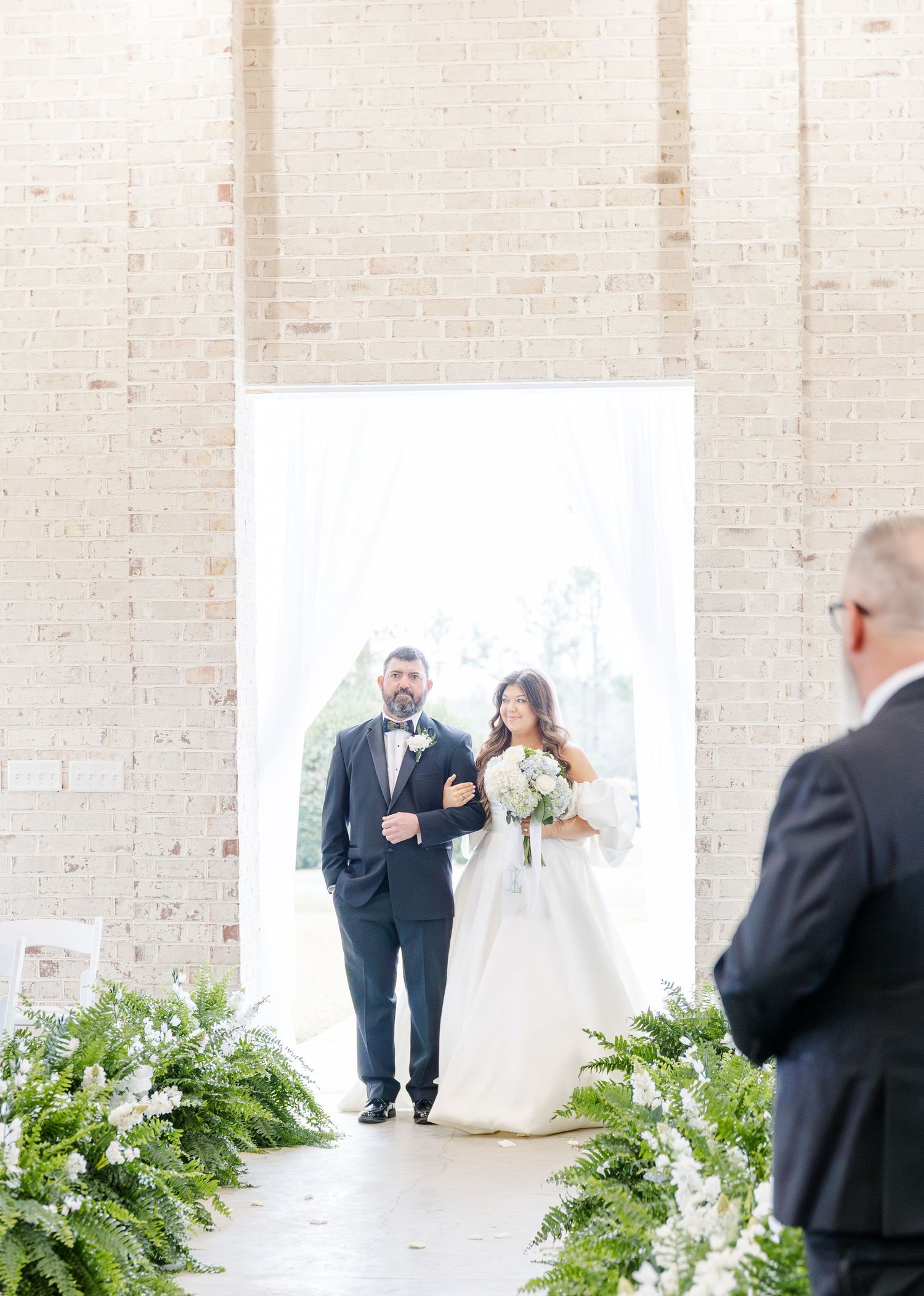 Bride walking down the aisle with her escort in a bright chapel lined with lush green ferns.