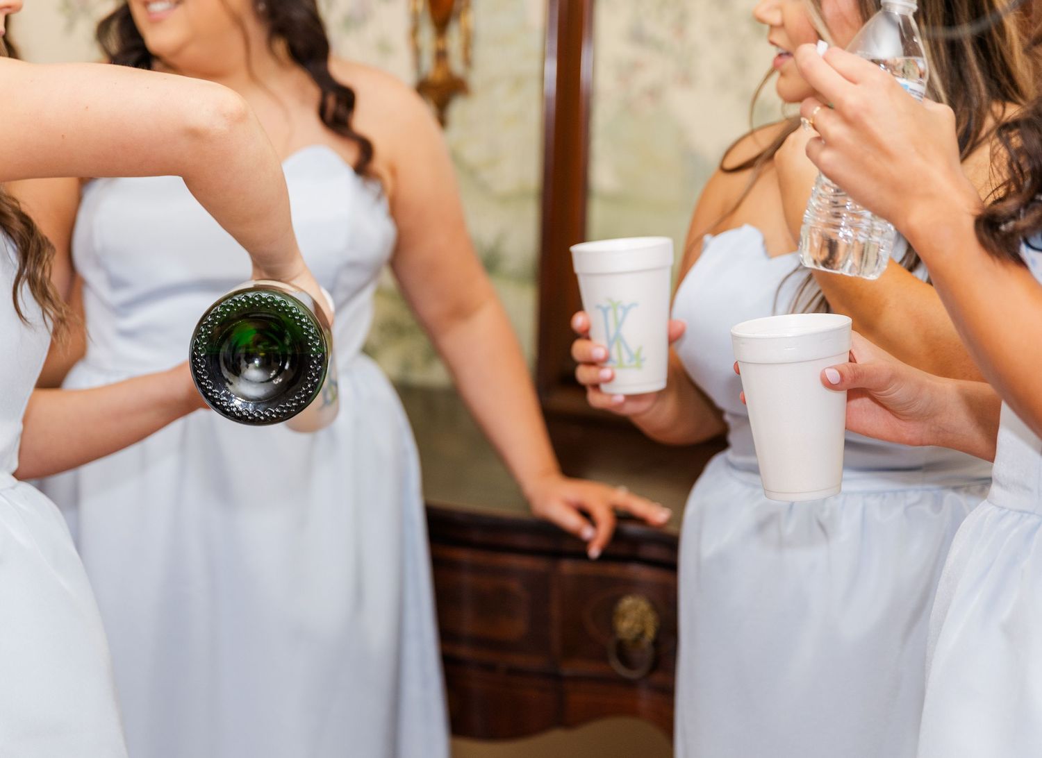 Bridesmaids in white dresses celebrating with champagne bottle and cups during wedding getting ready festivities.