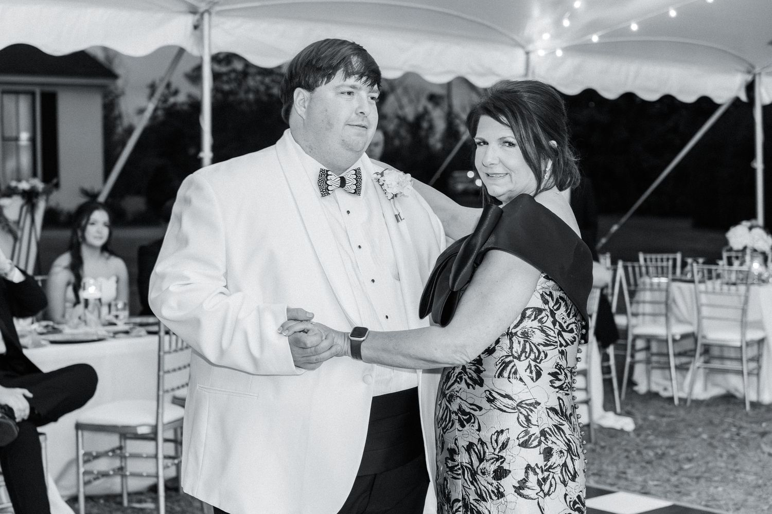Black and white photo of groom in white tuxedo dancing with woman in floral dress at reception.