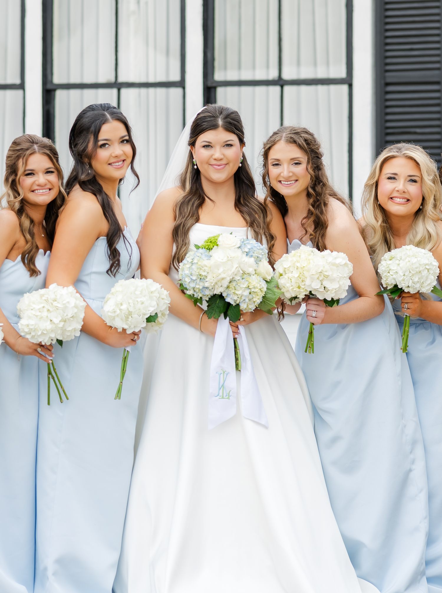 Bride in white gown poses with four bridesmaids in light blue dresses holding white hydrangea bouquets.