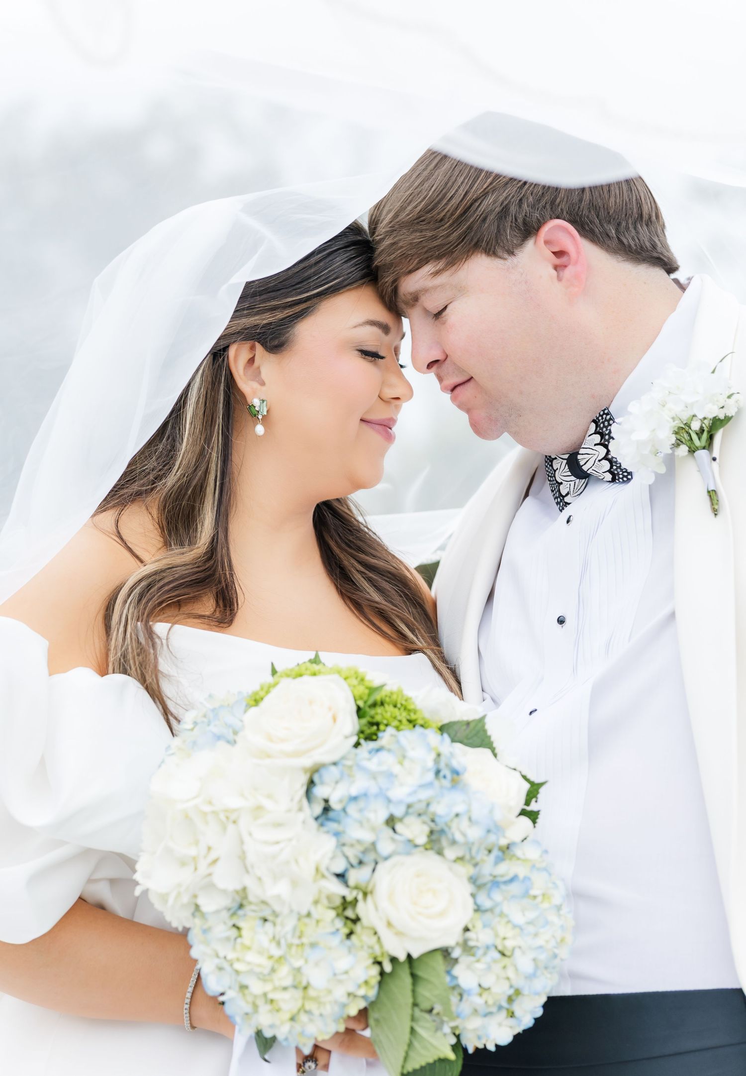Romantic bride and groom touching foreheads under veil, bride holding blue and white bouquet.