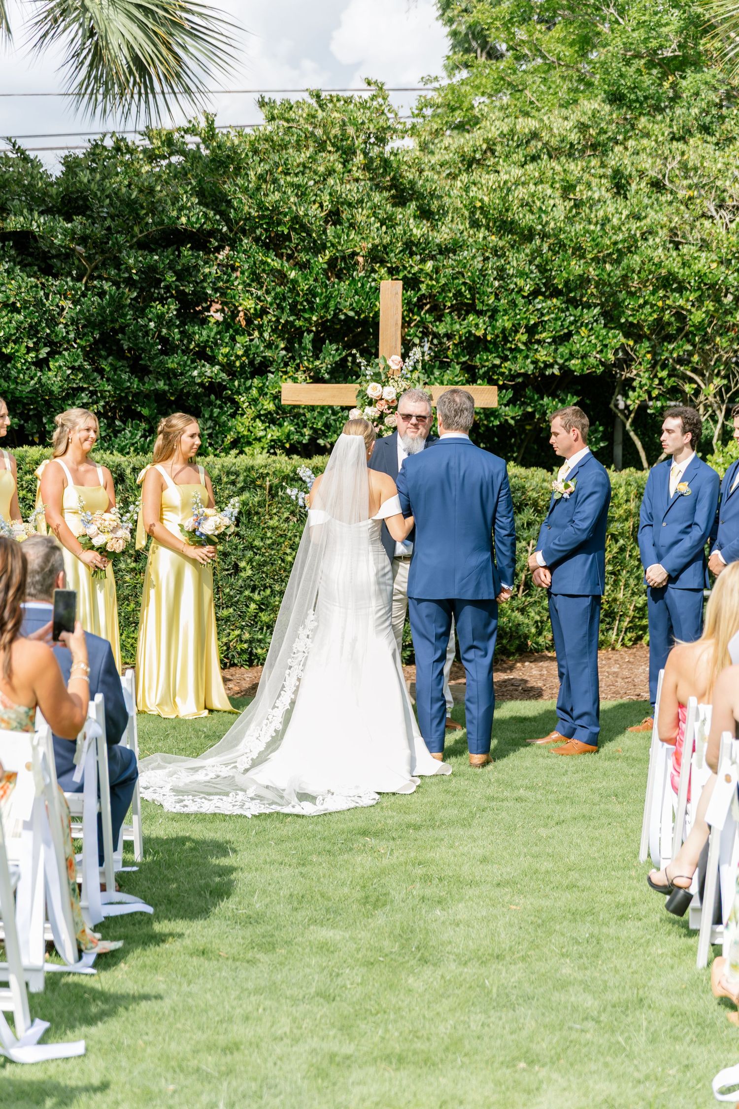 Outdoor wedding ceremony setup on a green lawn with white chairs, blue-suited groomsmen and yellow bridesmaid dresses.