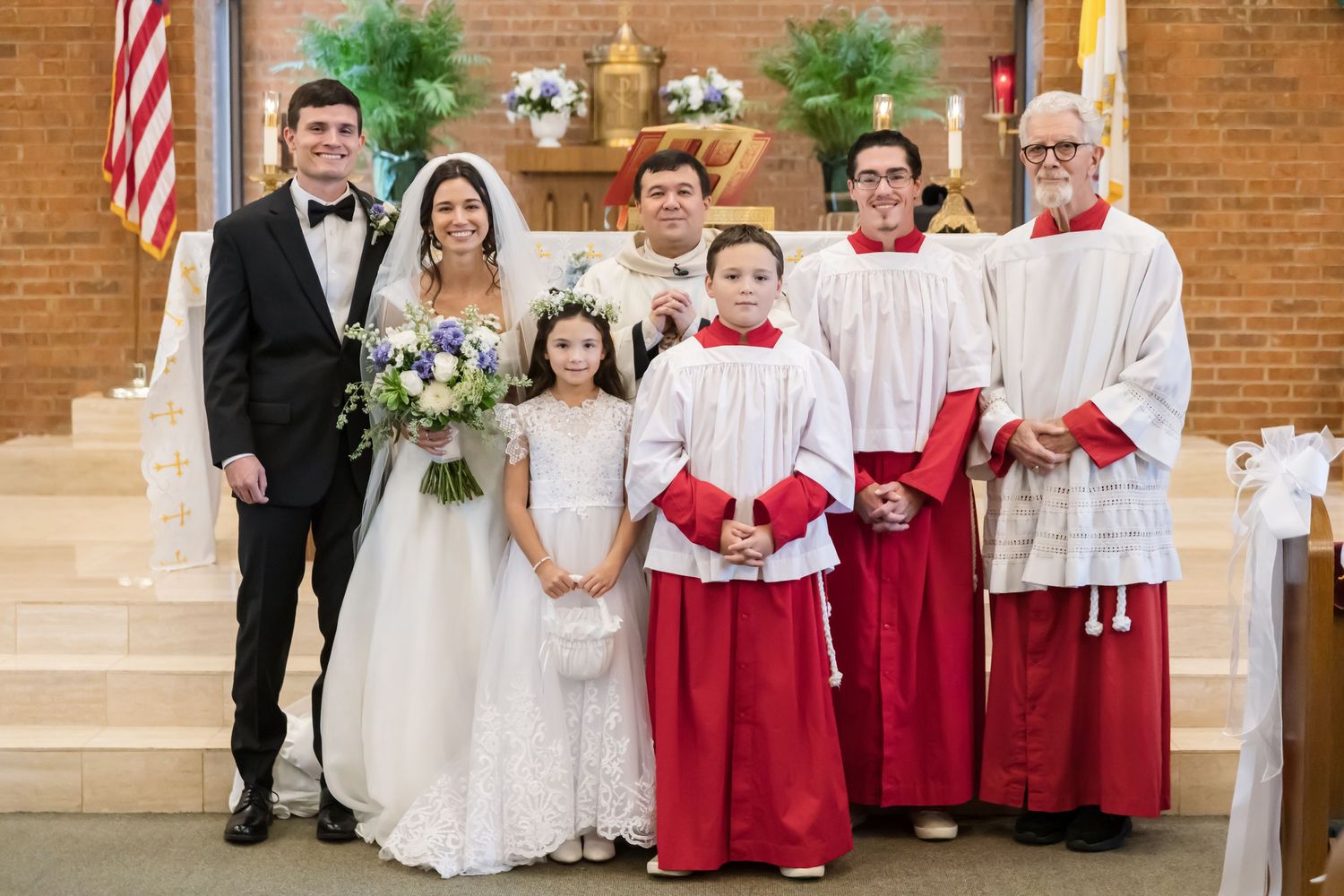 Wedding party members in formal attire stand together with choir members in red and white robes at a church ceremony.