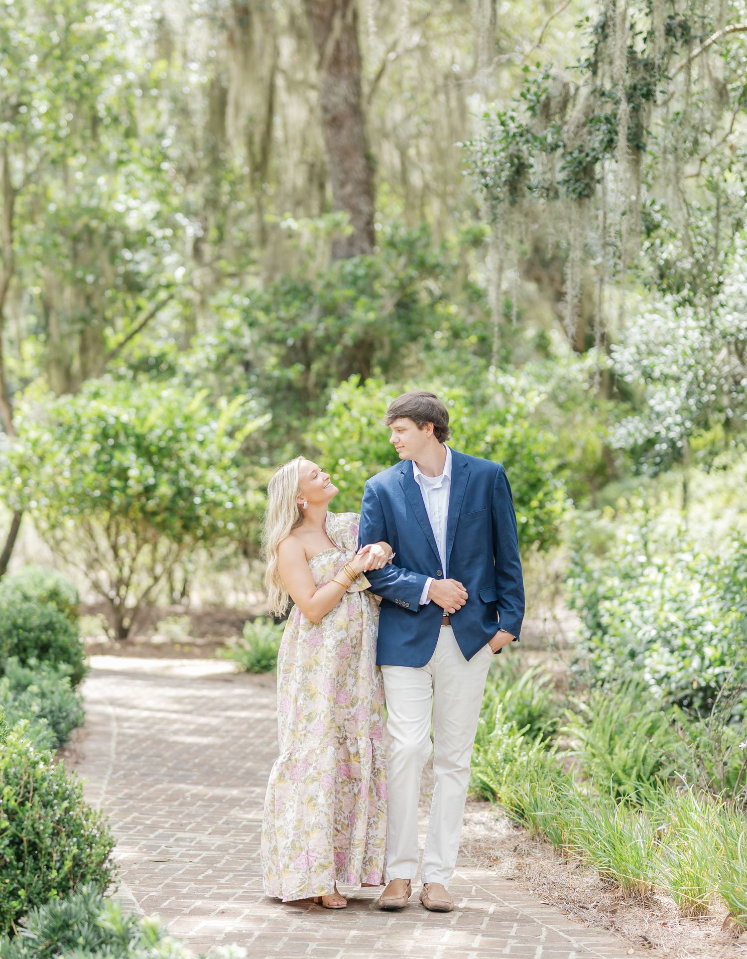 A couple walks together on a brick path surrounded by lush greenery and Spanish moss hanging from trees.
