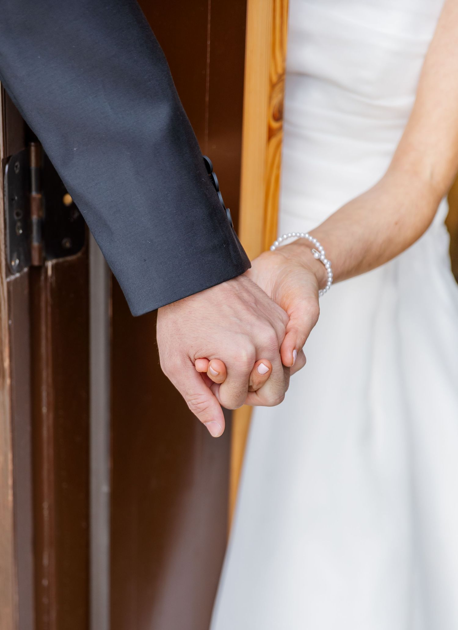 A couple in wedding attire holding hands, showing a delicate bracelet and formal sleeves in an intimate moment.