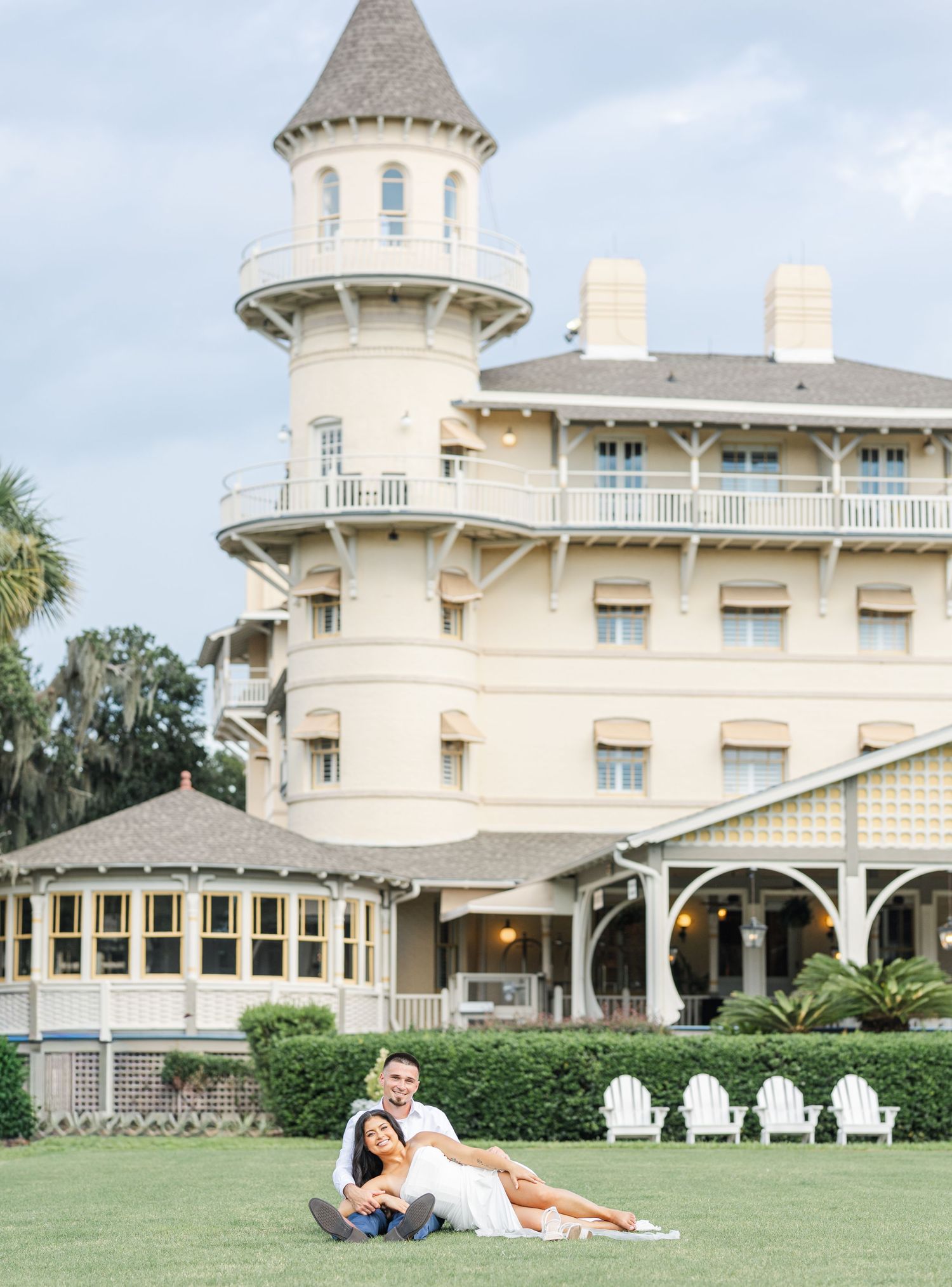 Couple sitting together on grass before grand yellow Victorian building with distinctive turret tower.