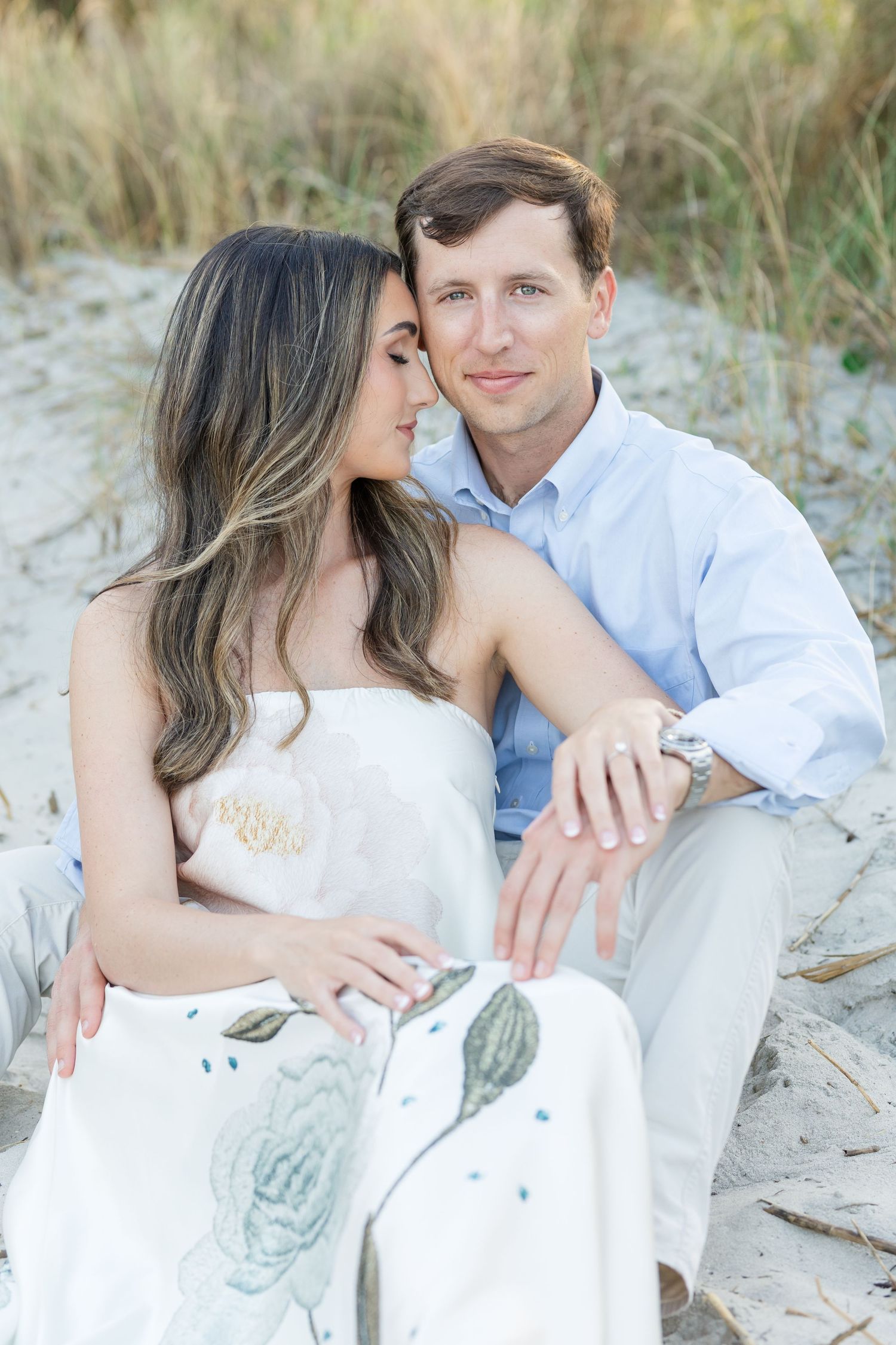 Couple sitting on sandy beach dunes, woman in floral white dress leaning on man in light blue shirt.