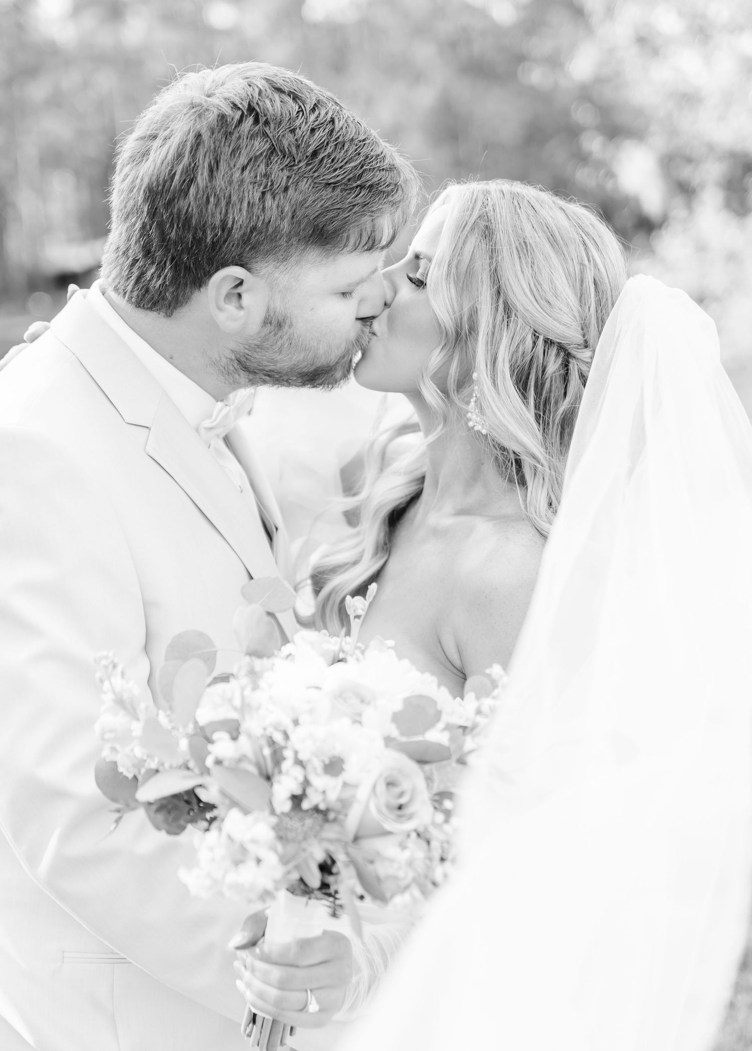 Black and white wedding photograph capturing an intimate kiss between bride and groom with delicate bouquet.
