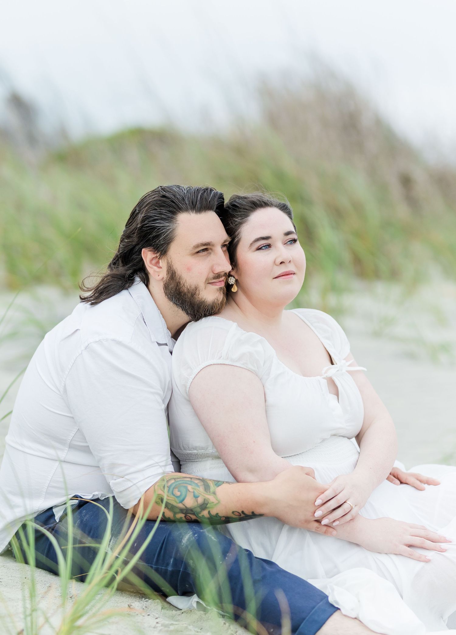 A romantic couple sits together on a sandy beach with tall grass in the background.
