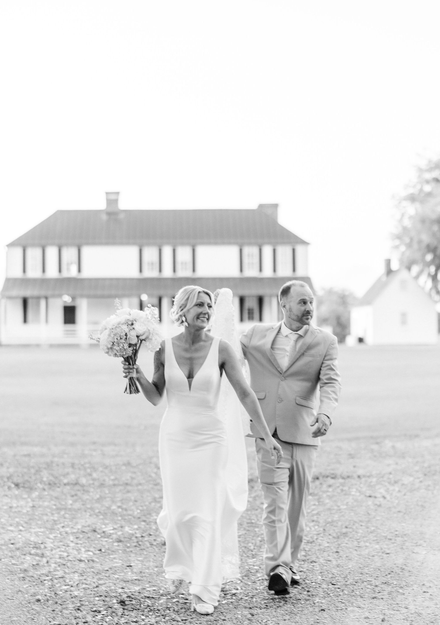 Black and white photo of bride holding bouquet and groom walking hand in hand near a historic white farmhouse.