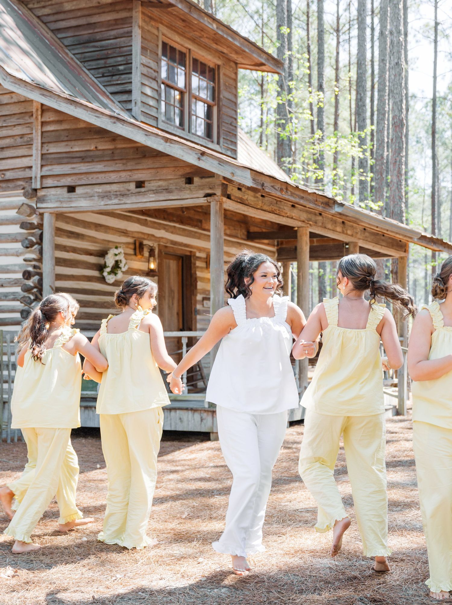 Bride in white holding hands with bridesmaids in yellow, dancing near a rustic log cabin wedding venue.