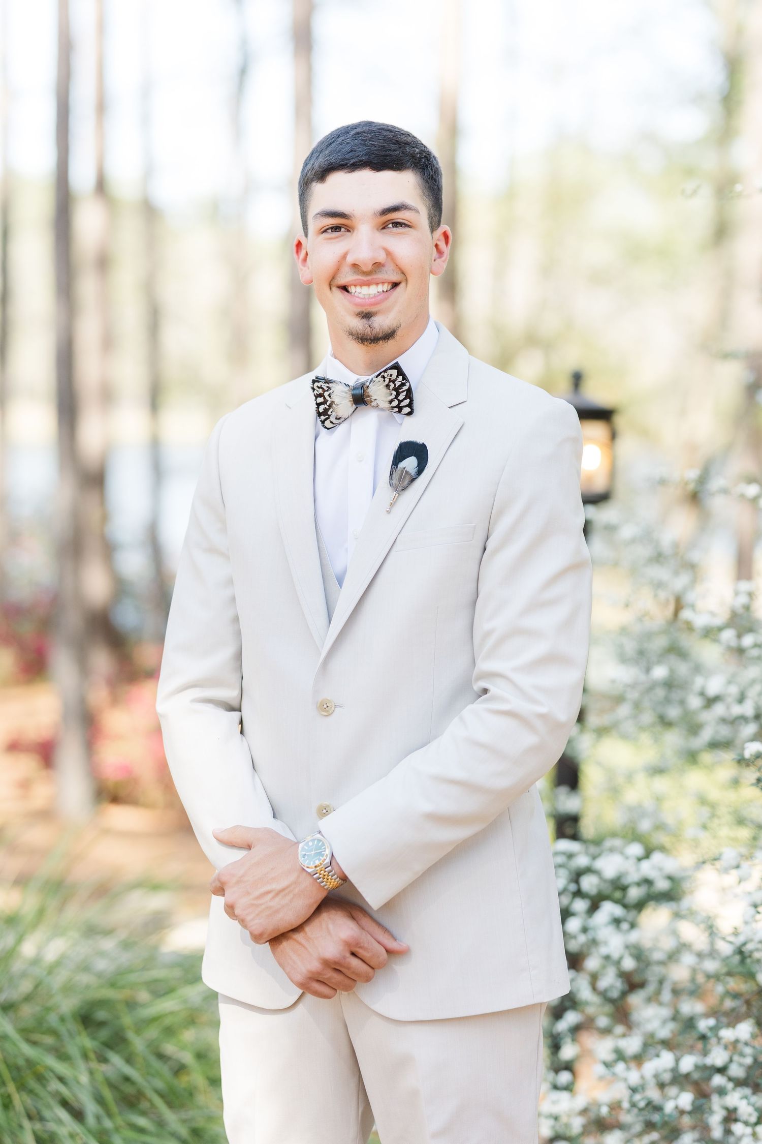 Smiling groom in ivory suit and patterned bow tie posing outdoors at wedding venue.