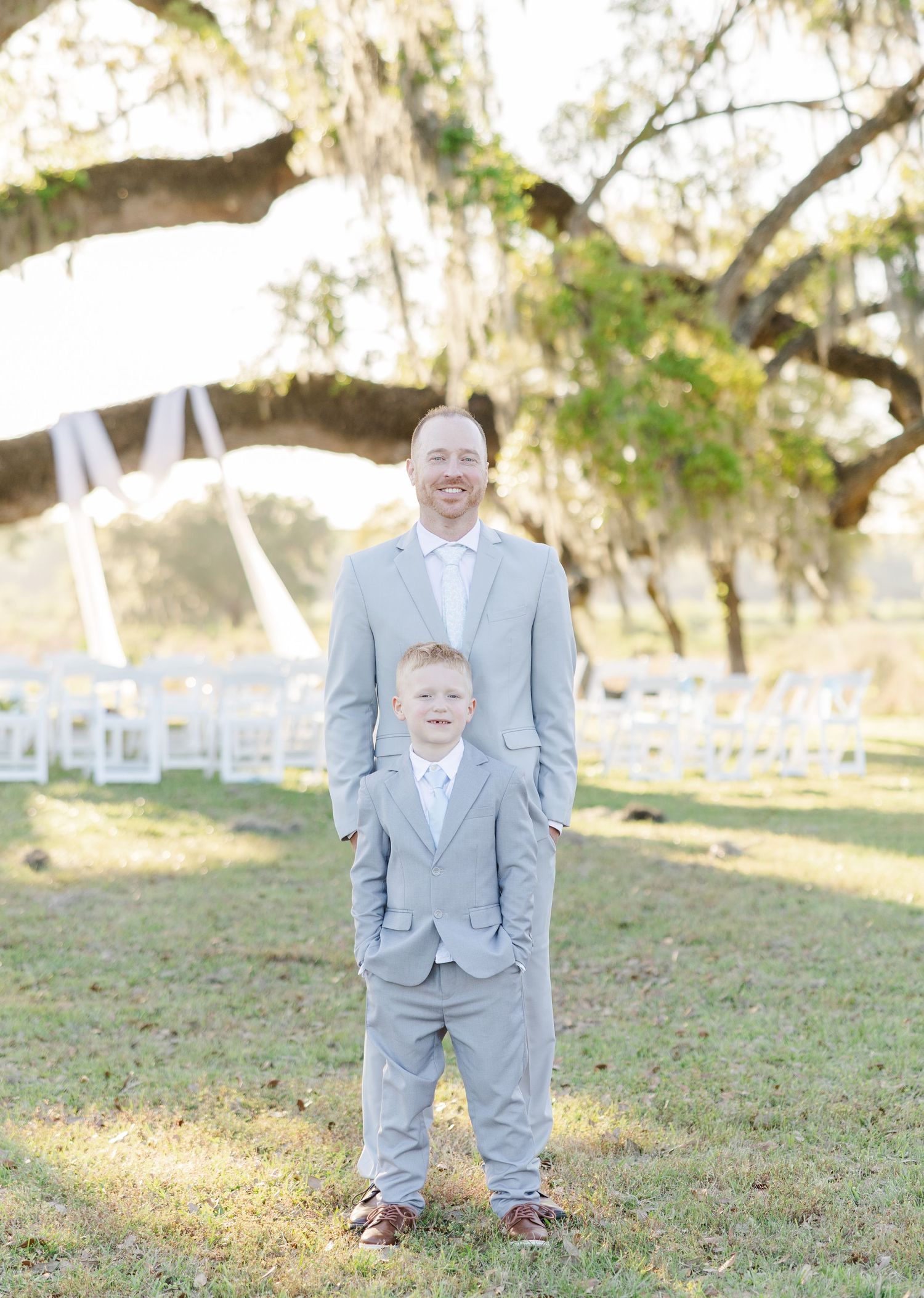 Groom in light gray suit poses with young ring bearer boy in matching suit under Spanish moss oak tree.